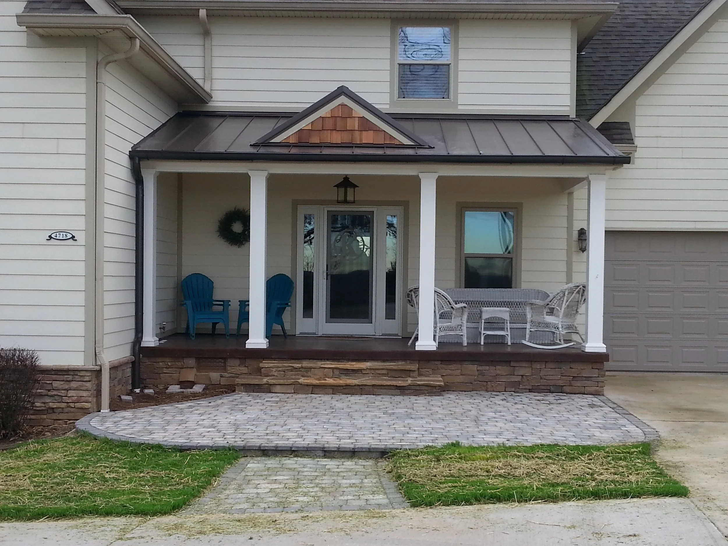 Front porch of a house with white siding, a metal roof, white columns, a glass door, a wreath, blue chairs, white wicker furniture, a hanging lantern, and a stamped concrete patio.