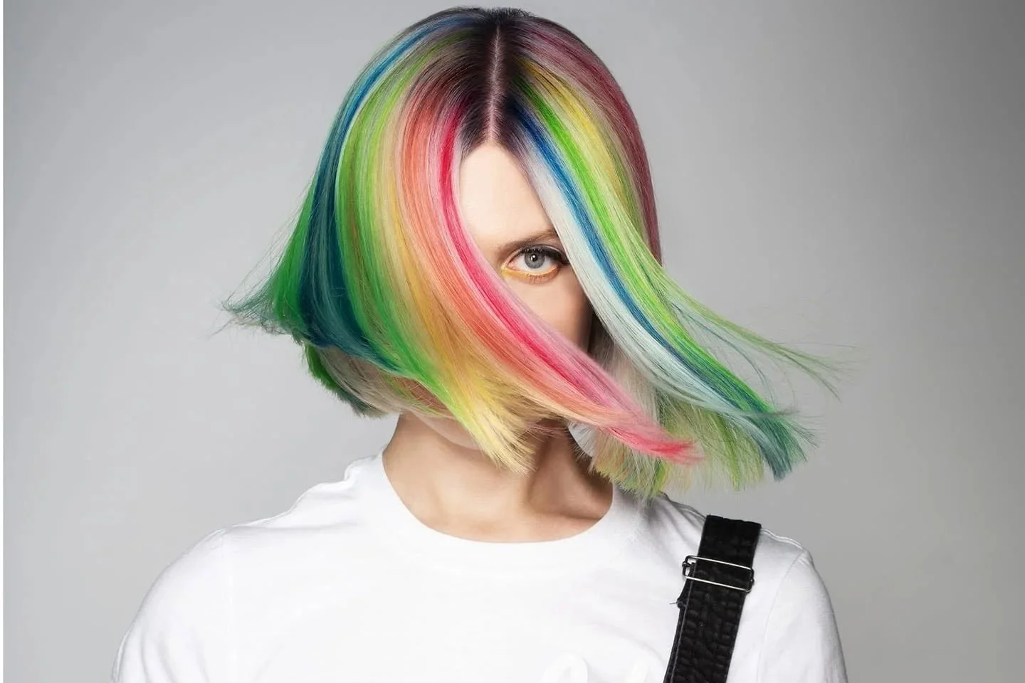 Woman with multicolored rainbow hair covering one eye, wearing a white shirt and black strap over shoulder, against a gray background.