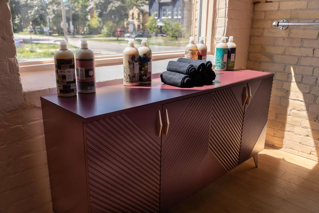 A pink sideboard with angled door panels and gold handles, topped with bottles of liquid soap or lotion and neatly folded black towels, positioned near a window with sunlight streaming in.