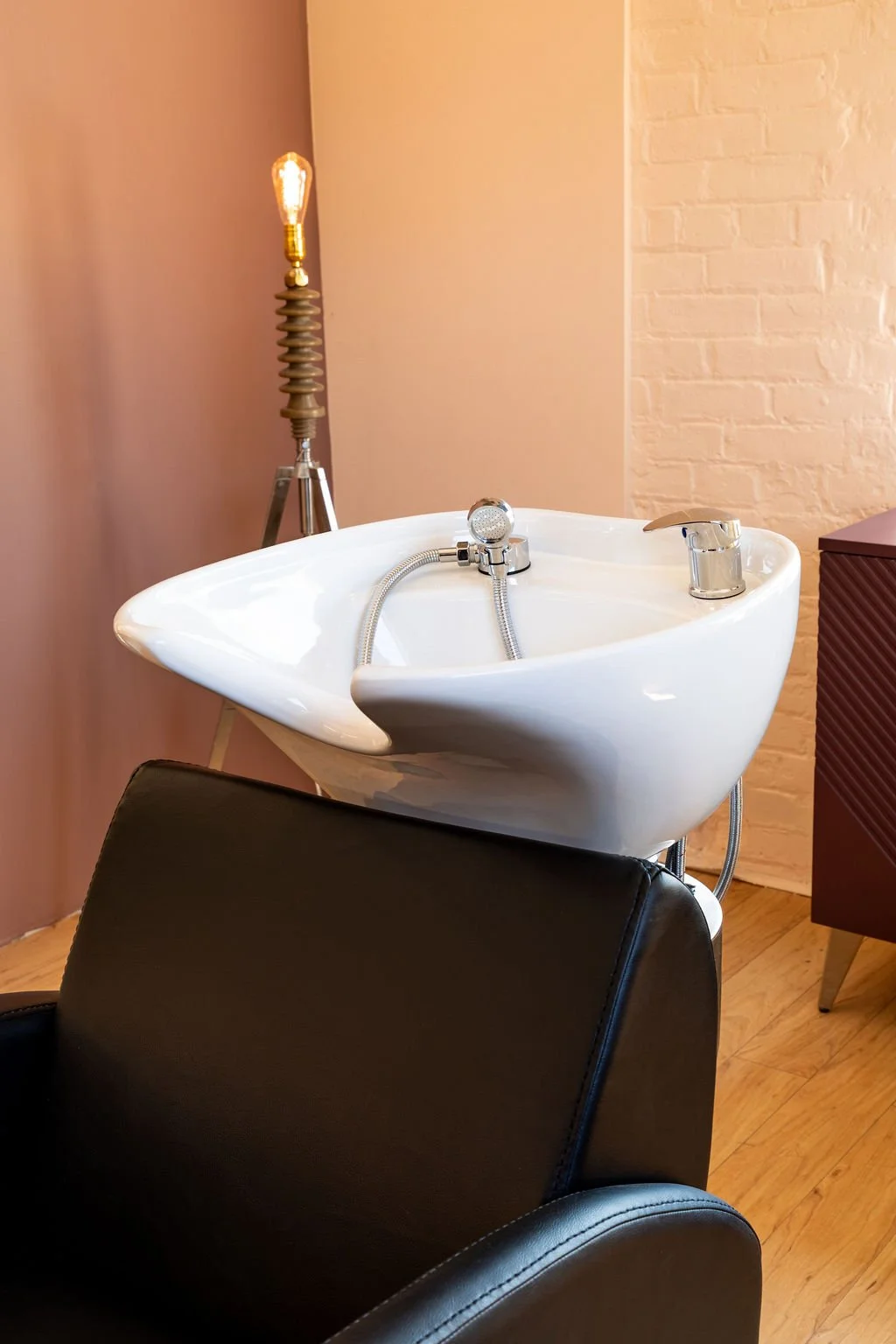 White salon shampoo bowl with taps, near a black chair, with a lamp in the background on a tripod stand.