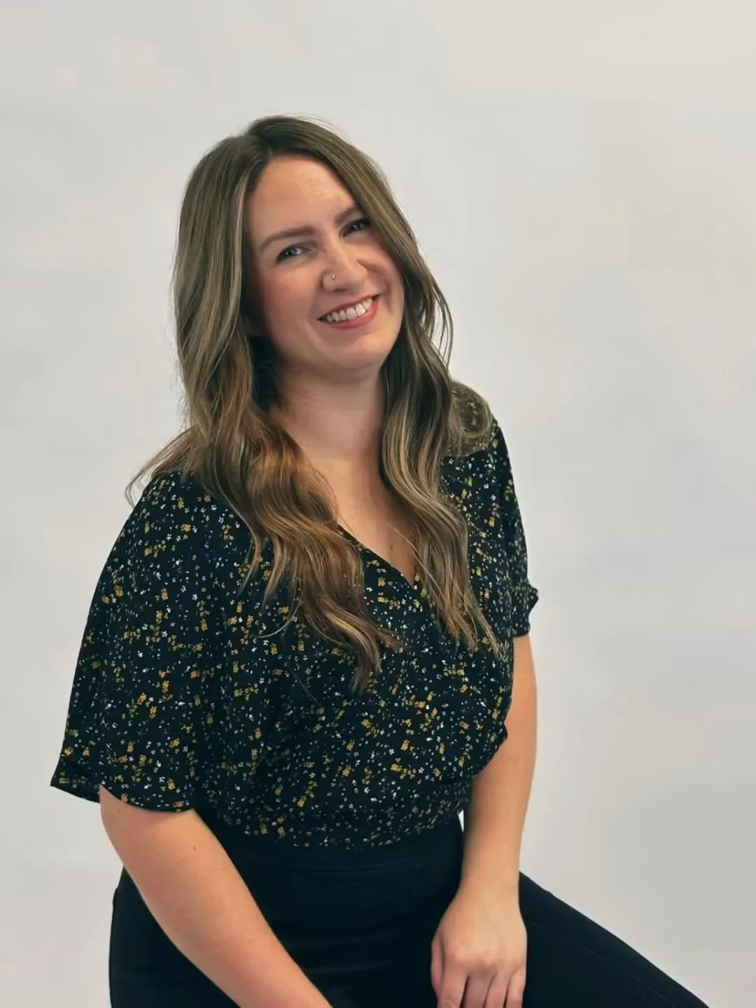 A young woman with wavy brown hair, a nose piercing, and a bright smile, wearing a black dress with yellow and white floral patterns, sitting against a plain white background.