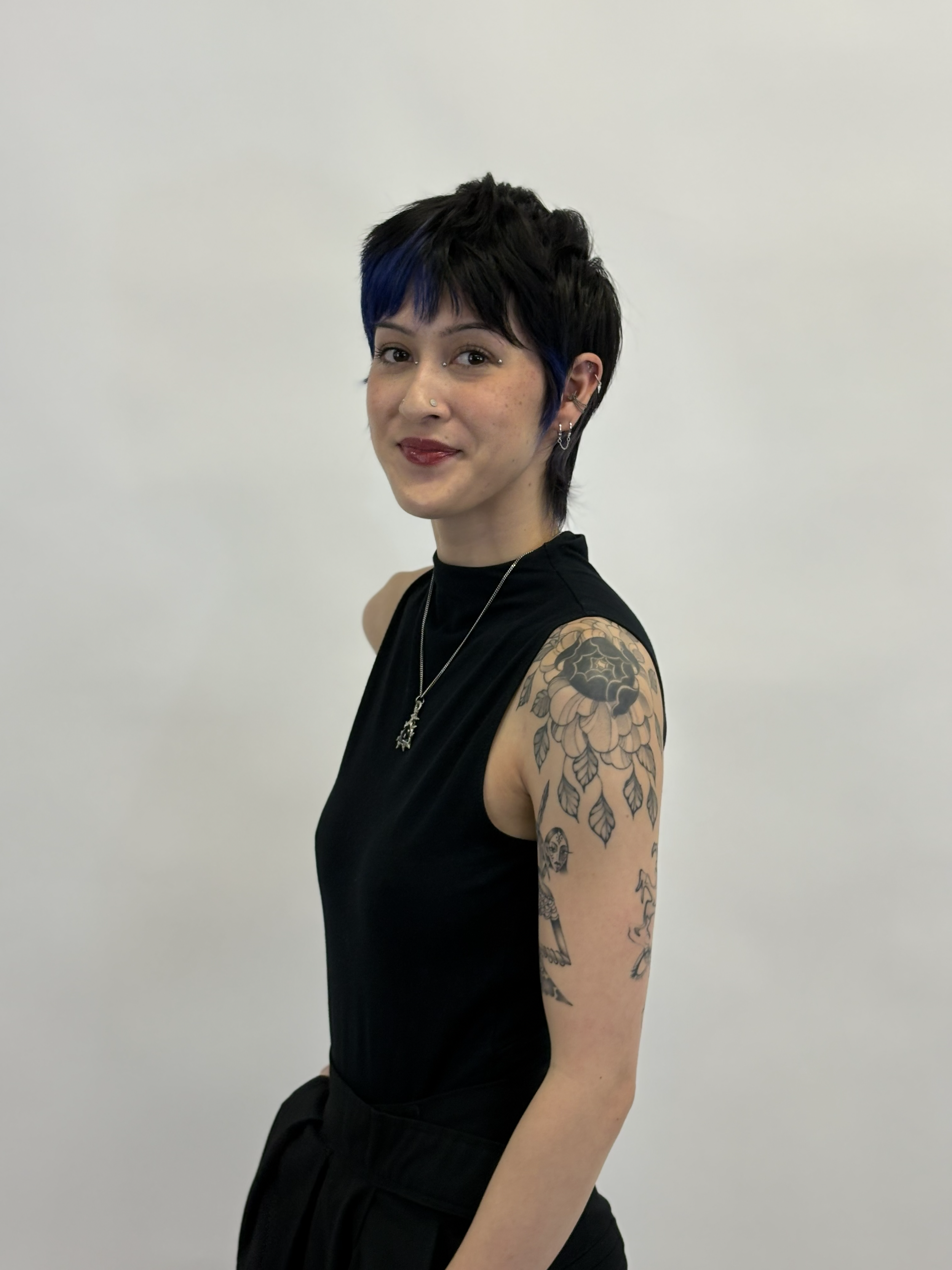 A woman with short black hair with blue highlights, wearing a black sleeveless top, showcasing tattoos on her left upper arm and shoulder, against a plain white background.