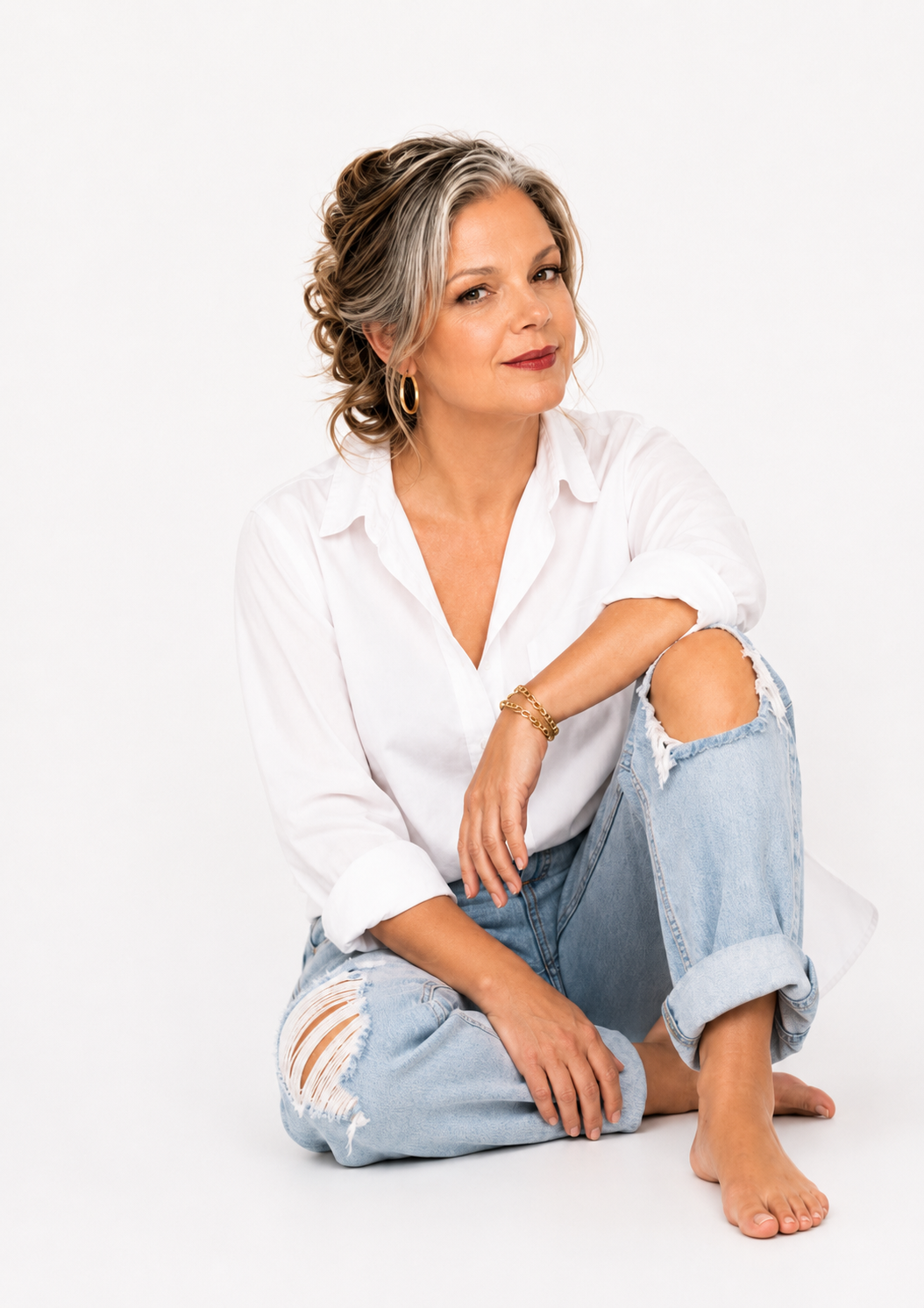 A mature woman with gray hair styled in loose curls, wearing a white button-up shirt with rolled-up sleeves, ripped jeans, gold hoop earrings, and a gold bracelet, sitting barefoot against a white background.