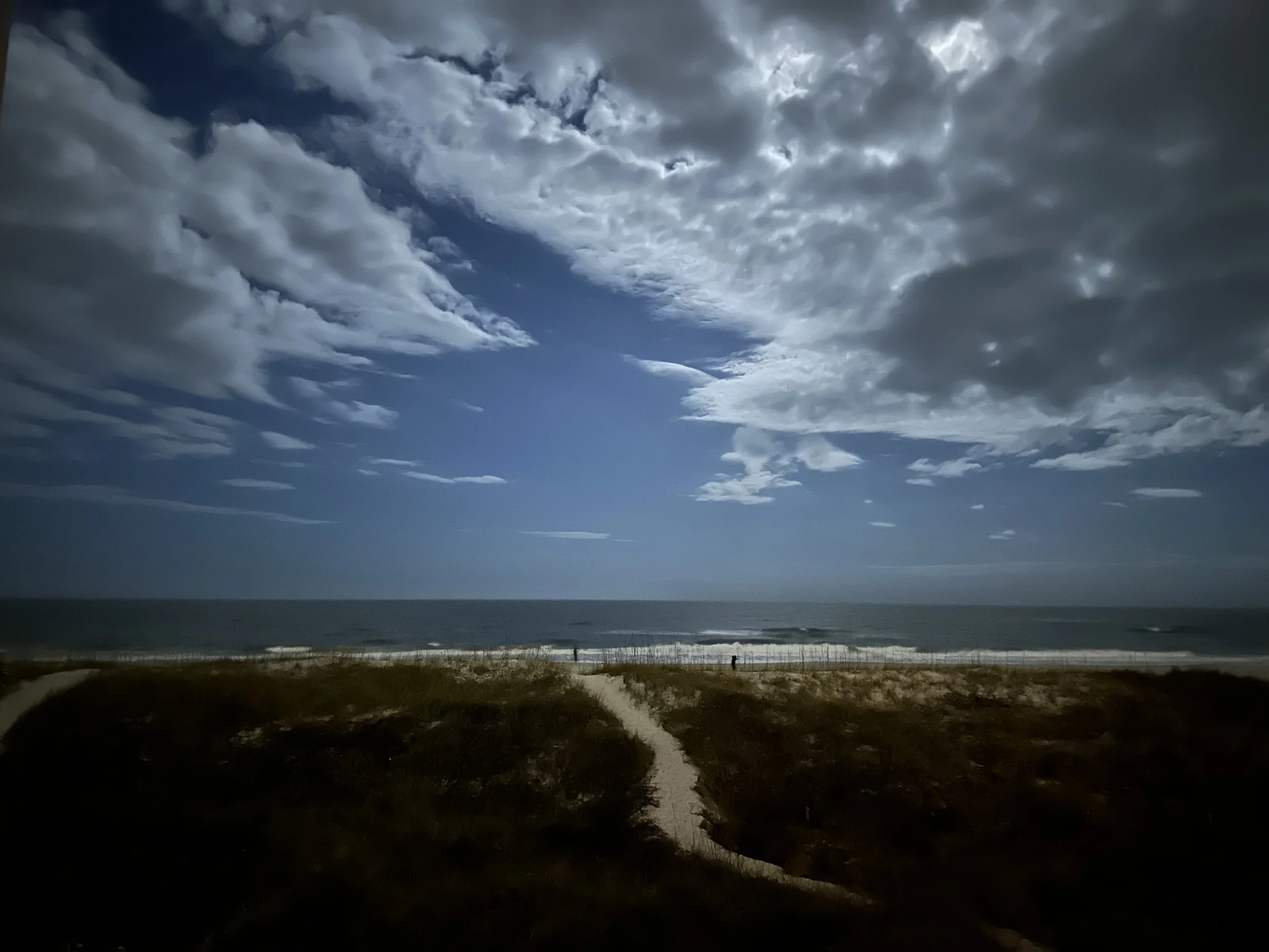 Cloudy sky over a beach with a single person walking along a sandy path bordered by dunes.