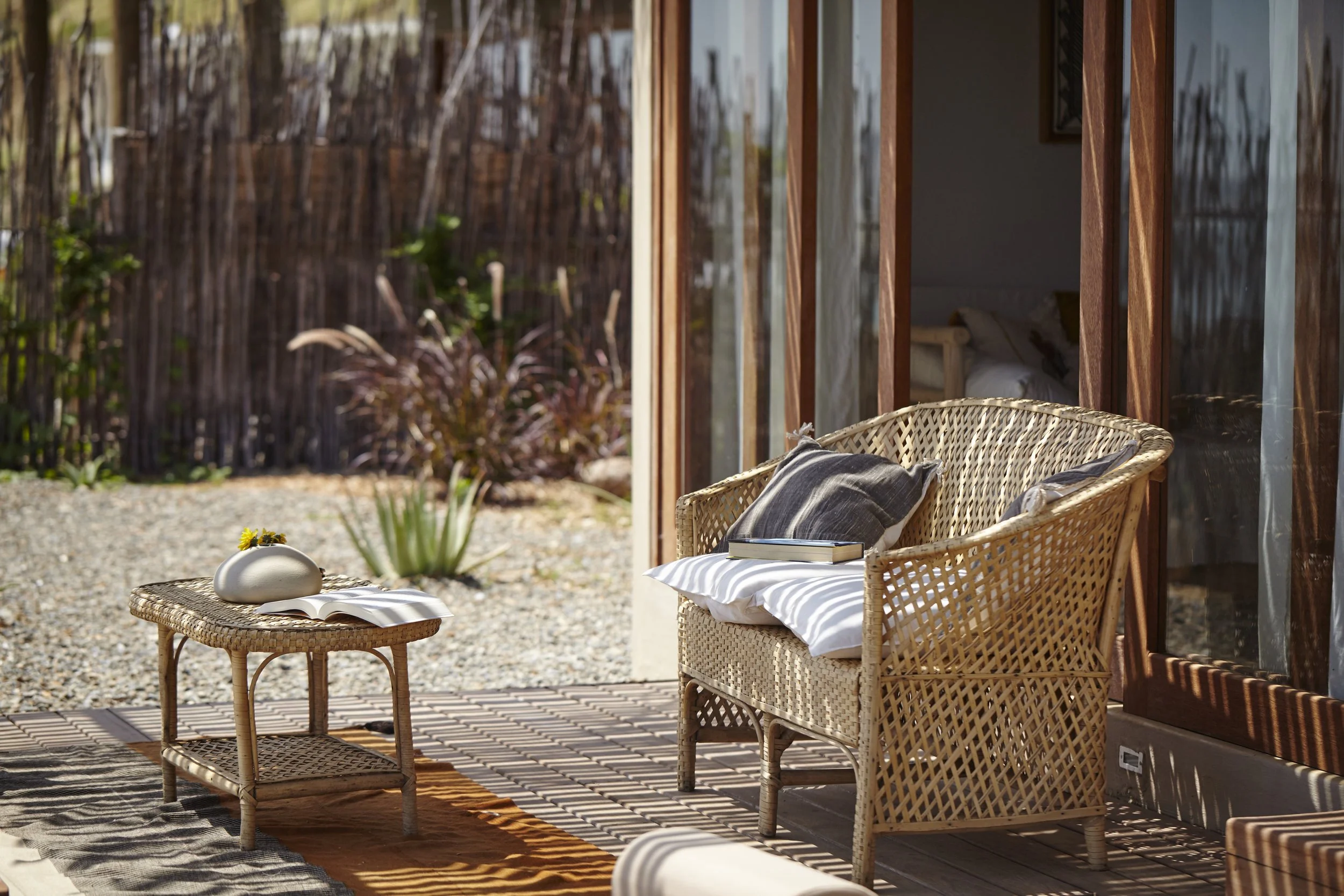Outdoor patio with wicker chairs, a small table with a book and decorative objects, and lush garden plants with a tall wooden fence in the background.