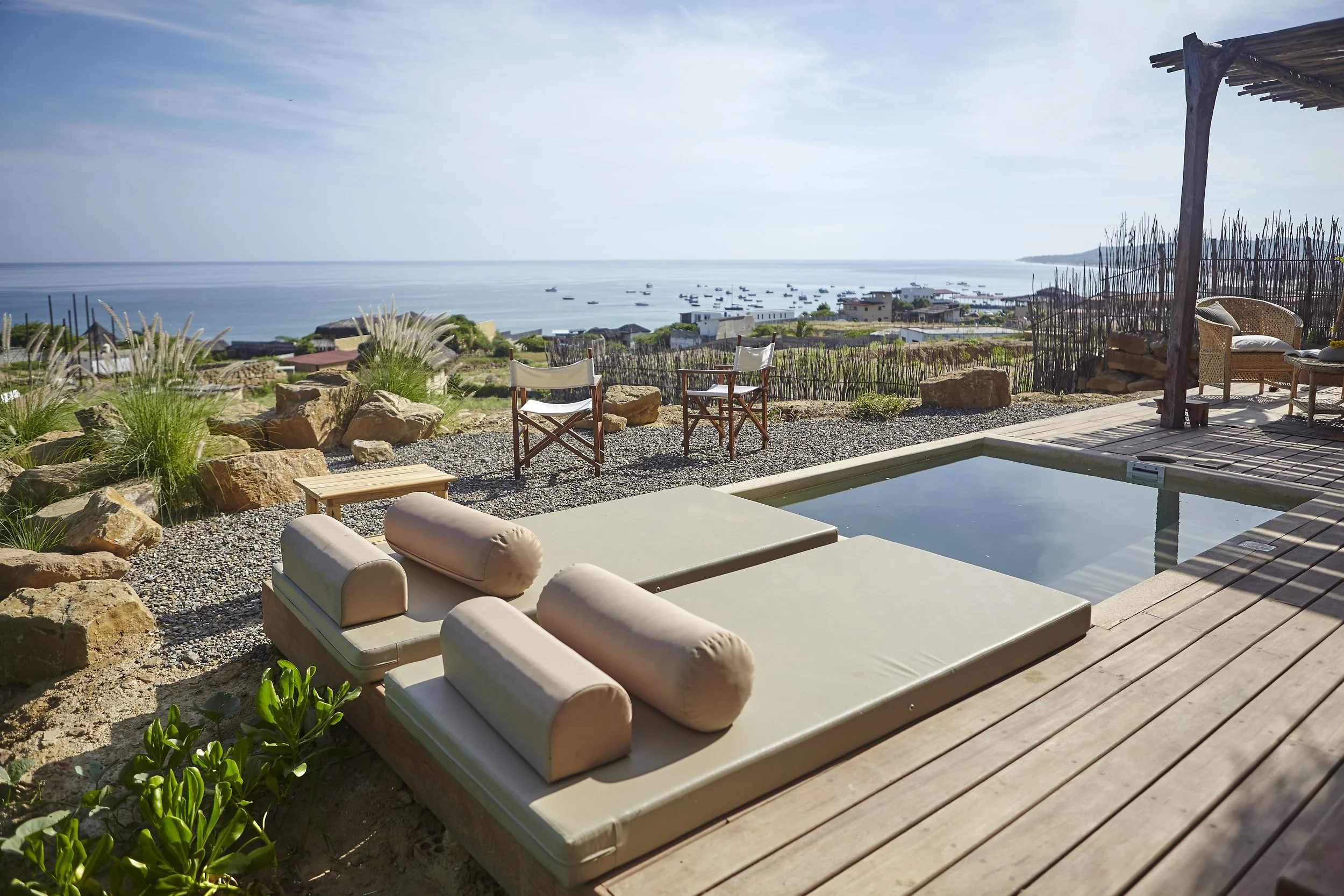 A scenic view of a coastal area from a patio with outdoor furniture, including cushioned loungers, a small table, wicker chairs, and a wooden deck, overlooking the ocean and boats on the water with a clear sky.