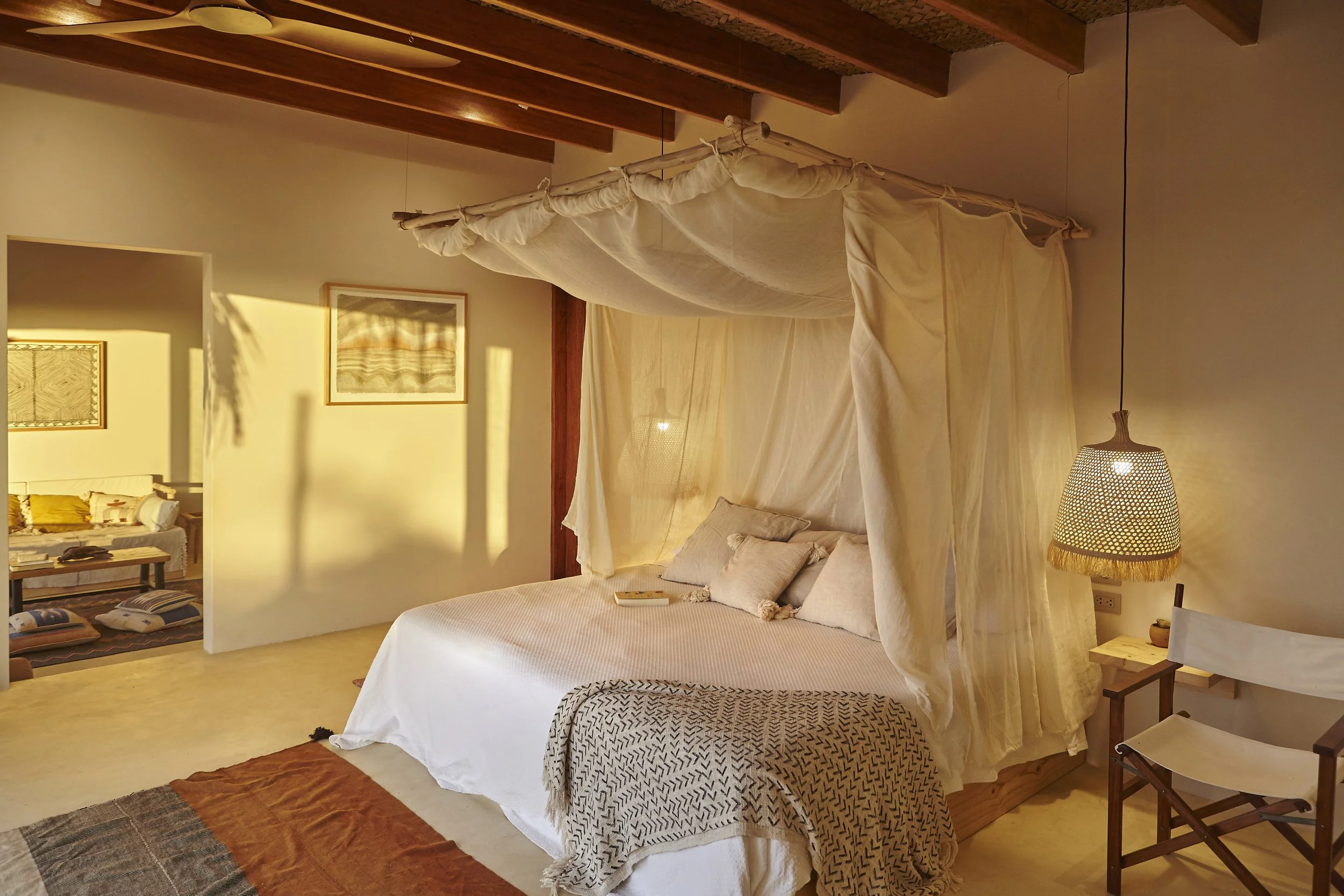 A cozy bedroom with a large bed featuring white linens, beige pillows, and a black-and-white patterned blanket. A canopy made of sheer, light-colored fabric hangs above the bed. To the right, there is a lamp with a woven, basket-like shade. The room 