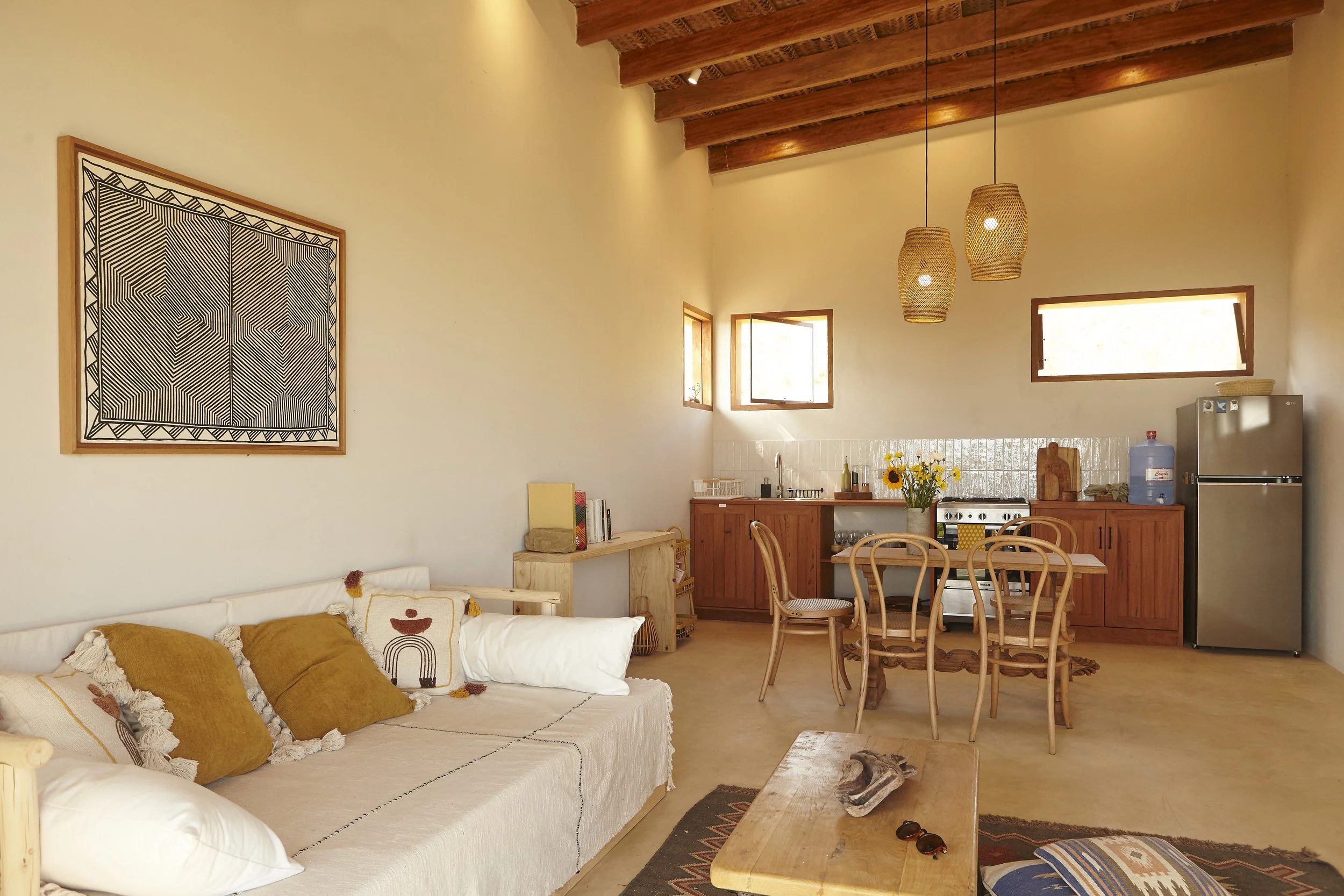 A cozy living space with a beige sofa decorated with mustard yellow and off-white pillows, a wooden dining table with chairs, and a kitchen area with wooden cabinets, small windows, and walls decorated with a black and white geometric art piece. The 