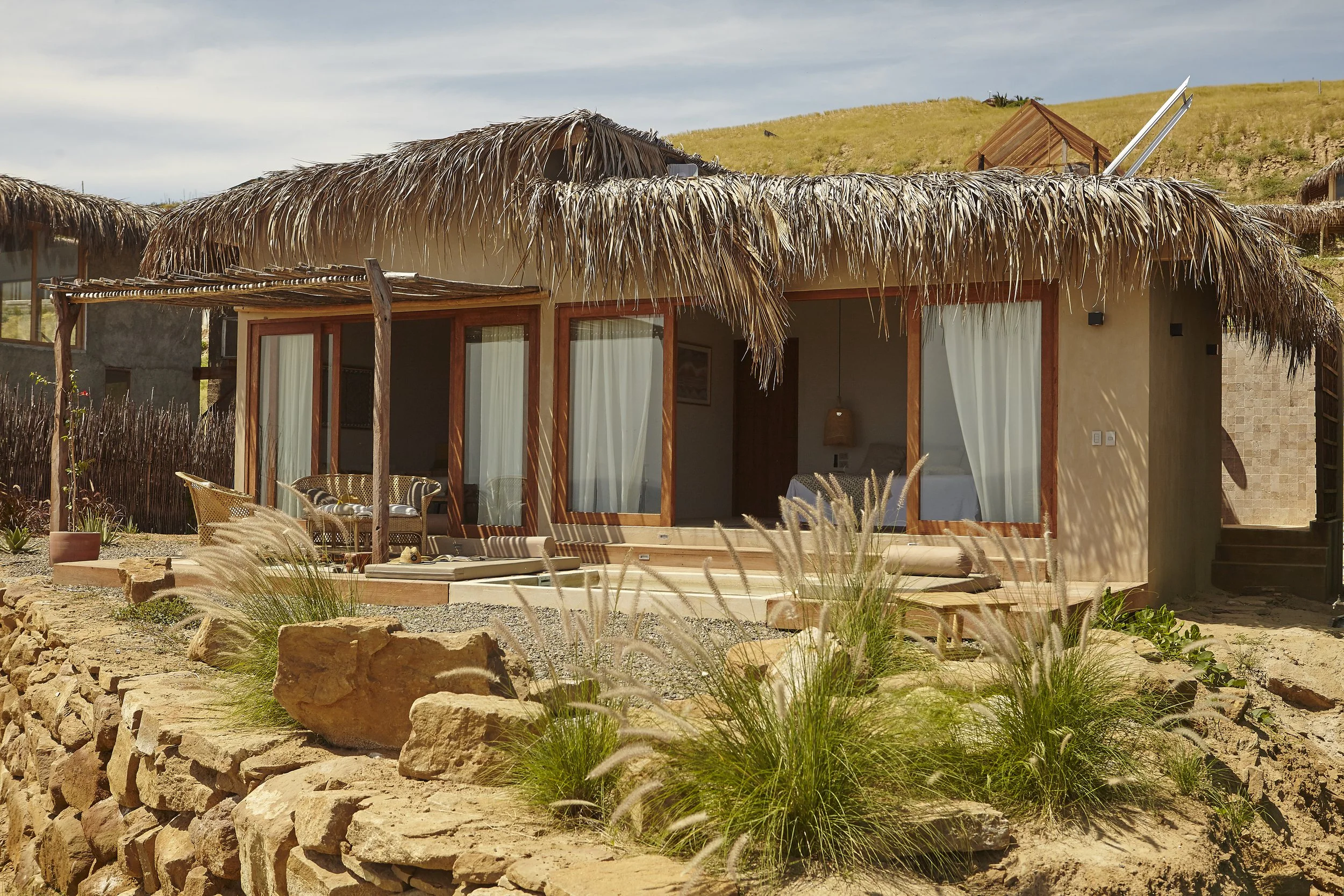 A thatched roof bungalow with large glass sliding doors, outdoor seating, and a stone and dirt yard with plants and rocks in a rural, hilly area.