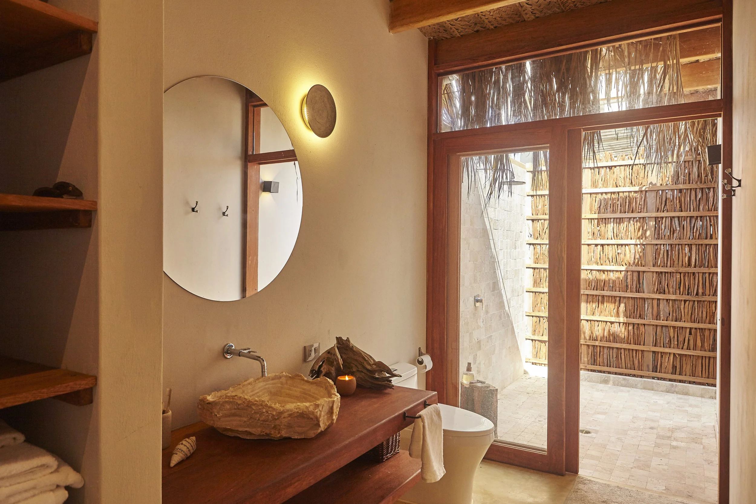 A bathroom with a large wooden-framed window and a thatched roof view outside. Inside, there is a rustic stone sink, wooden shelves with towels, a mirror, and a candle on the vanity.