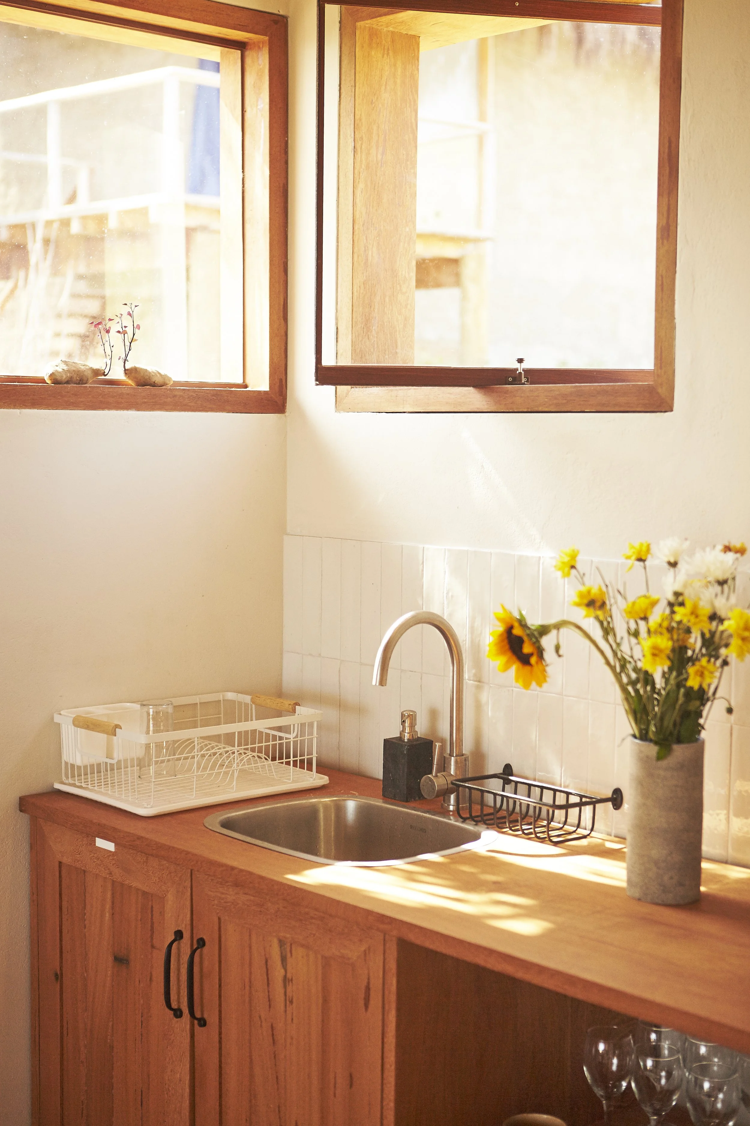 Kitchen with wooden cabinets, a sink, a window with sunlight coming through, a dish rack, a vase of yellow and white flowers, and glasses underneath.