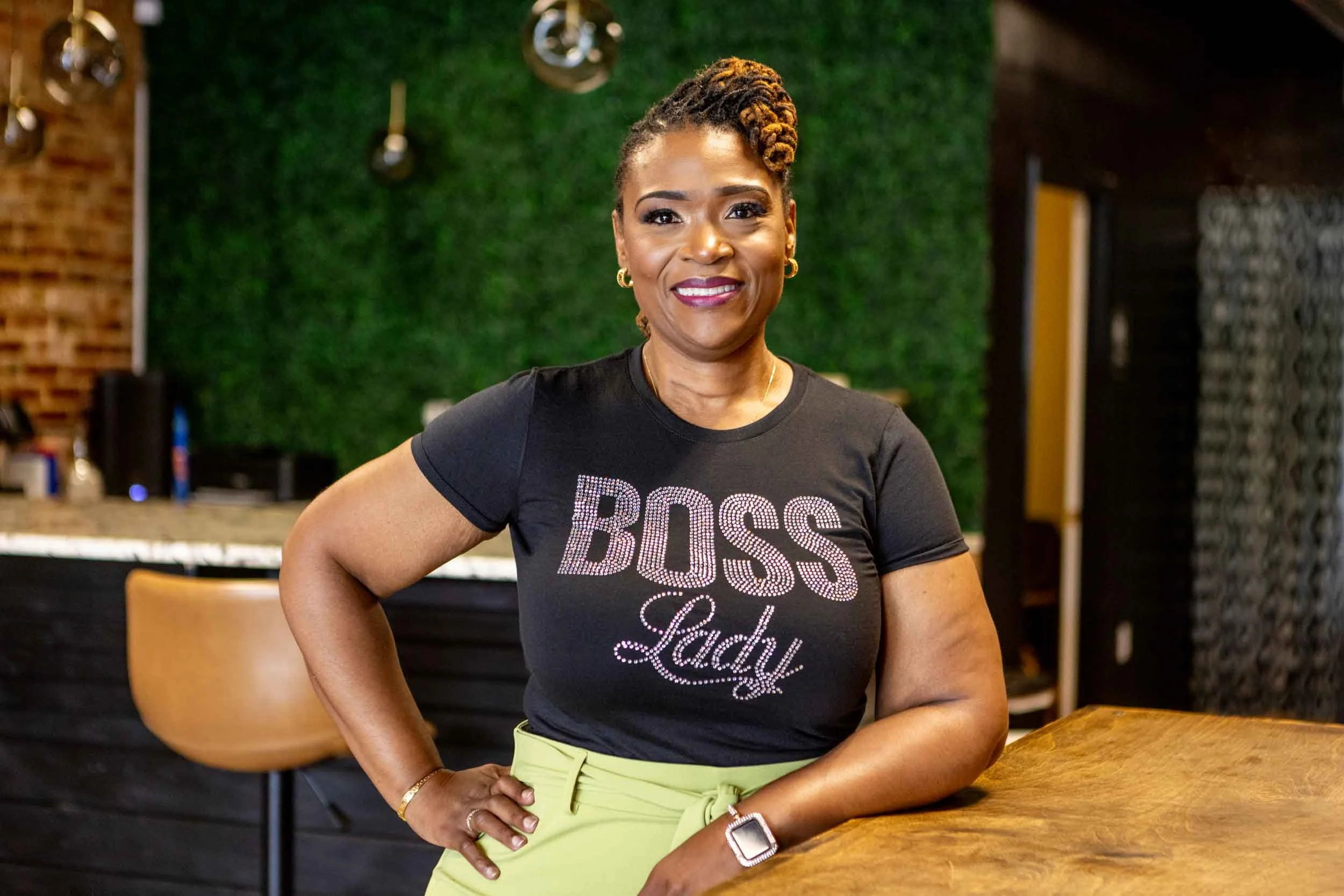 A woman with dark skin and curly hair styled into a side updo, smiling confidently, stands indoors in front of a green wall with a rustic brick wall and hanging lights in the background. She is wearing a black T-shirt with the words 'Boss Lady' in rhinestones, light green pants, a watch, and jewelry.