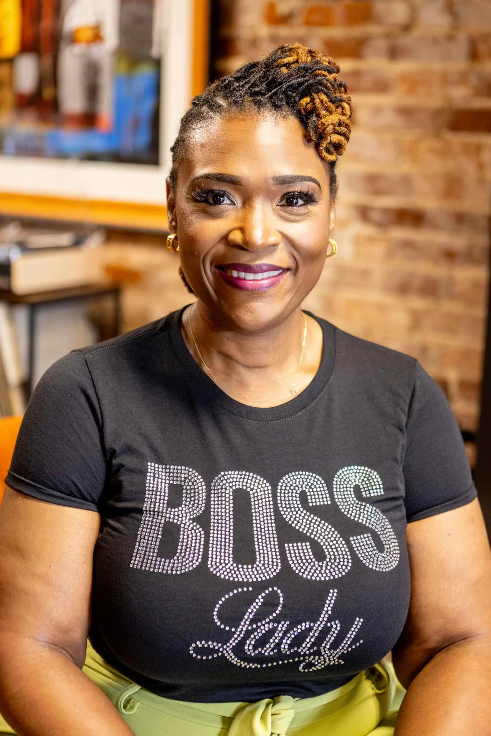 Close-up of a smiling Black woman with styled dreadlocks, wearing a black T-shirt with the word 'BOSS' in rhinestones and gold jewelry, sitting in front of a brick wall.