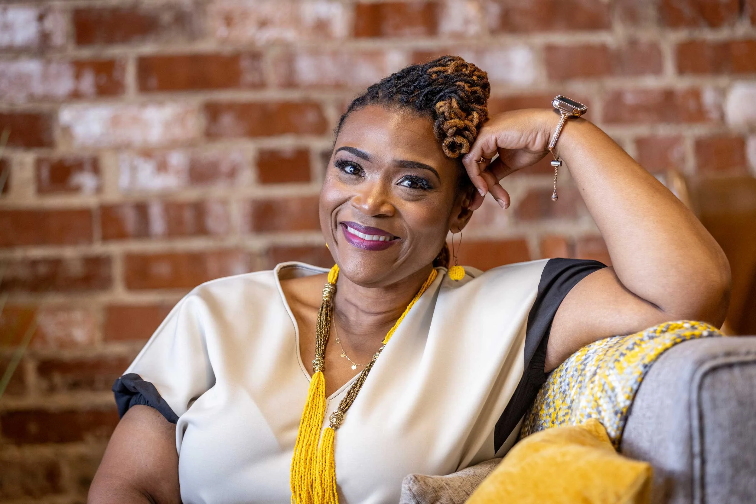 A smiling woman with twisted hair, wearing a beige top with black accents, yellow jewelry, and a watch, sitting against a brick wall.