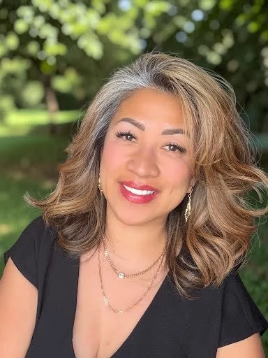 A woman with shoulder-length hair outdoors, smiling with green foliage in the background.