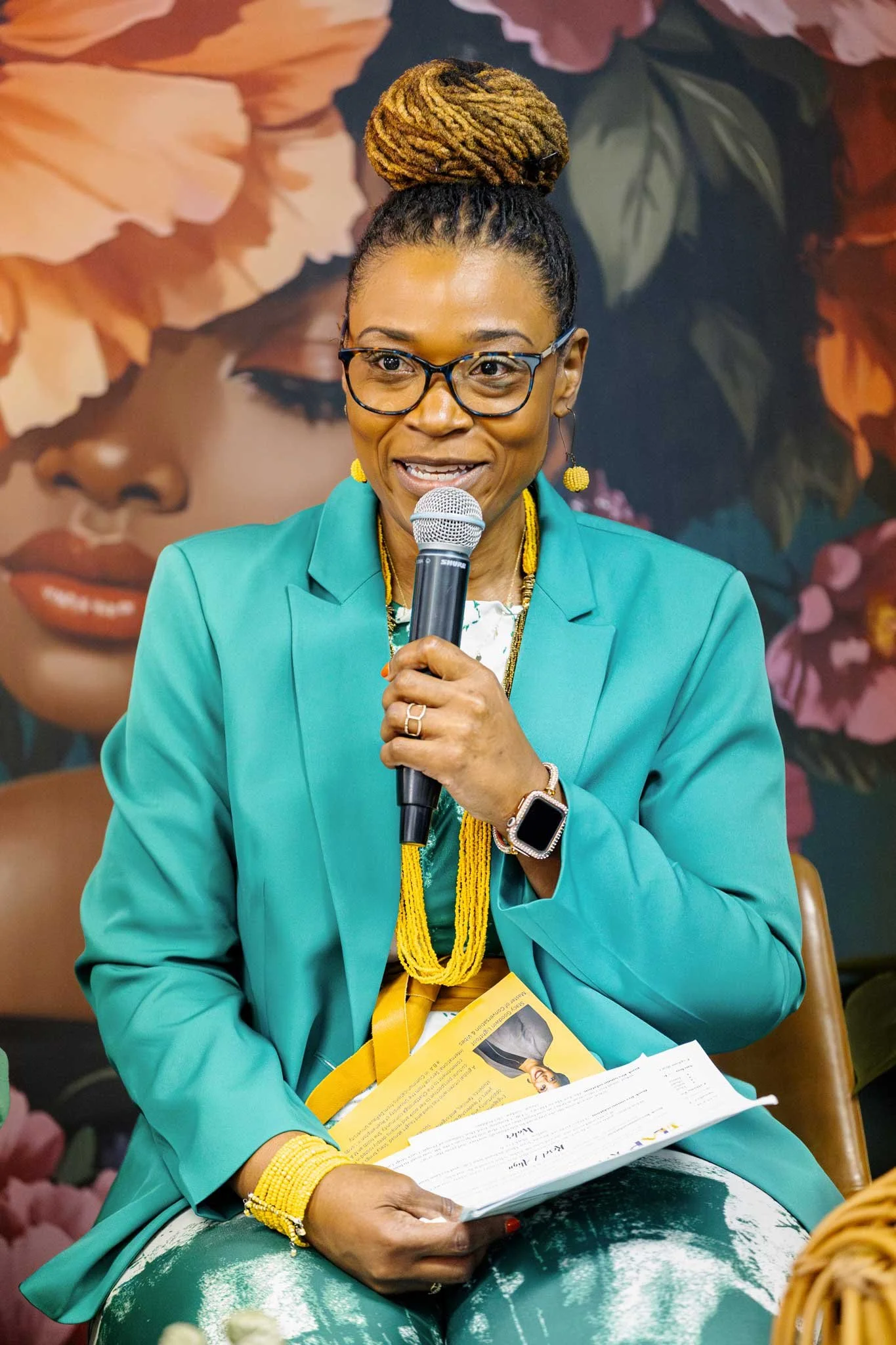 A woman with glasses and styled hair in a high bun is speaking into a microphone while sitting at a table. She is wearing a teal blazer, yellow jewelry, and a smartwatch. She is holding and looking at informational papers. Behind her, there is a large floral artwork of a woman's face.