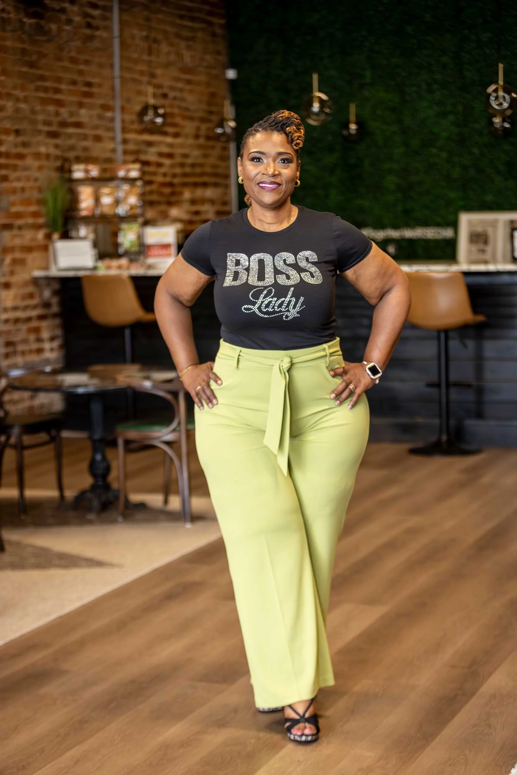 A woman standing with her hands on her hips in a cafe or restaurant with brick walls, green plants, and chairs in the background. She is wearing a black t-shirt with the words 'BOSS Lady' and yellow pants.