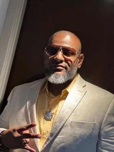 A man with a beard and sunglasses wearing a tan suit jacket, yellow shirt, and a necklace with a pendant. He is making a peace sign with his right hand, standing in front of a dark background.