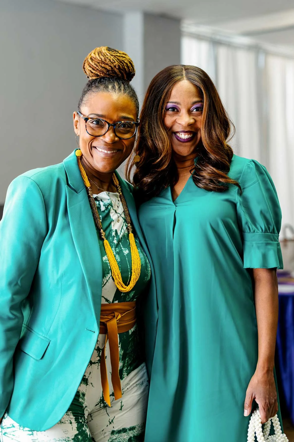 Two women smiling and posing together indoors, dressed in teal-colored outfits, with one wearing glasses, jewelry, and a hair bun, and the other with long, wavy hair and purple makeup.