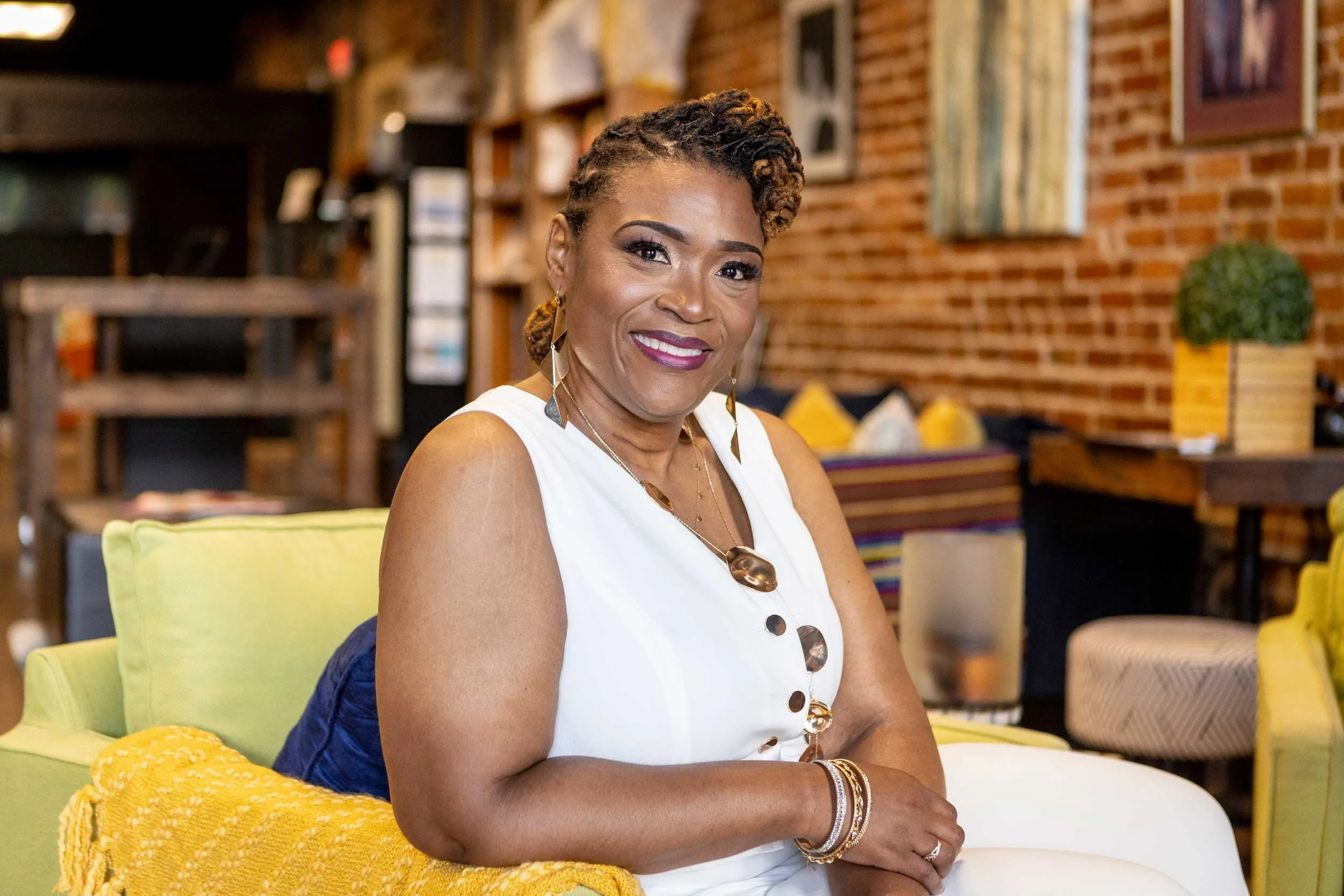 A woman with short, curly hair and dark skin is sitting on a yellow sofa in a cozy, modern living room. She is smiling and wearing a white sleeveless top with large, decorative buttons and gold jewelry. The background features a brick wall, framed artwork, and various decorative items.