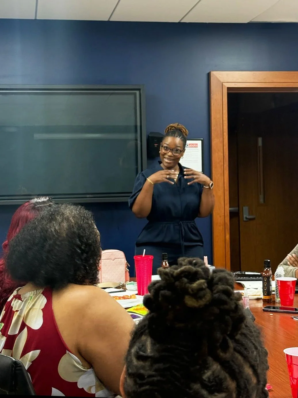 A woman standing and speaking to a seated group in a conference room, with others listening and several drinks on the table.