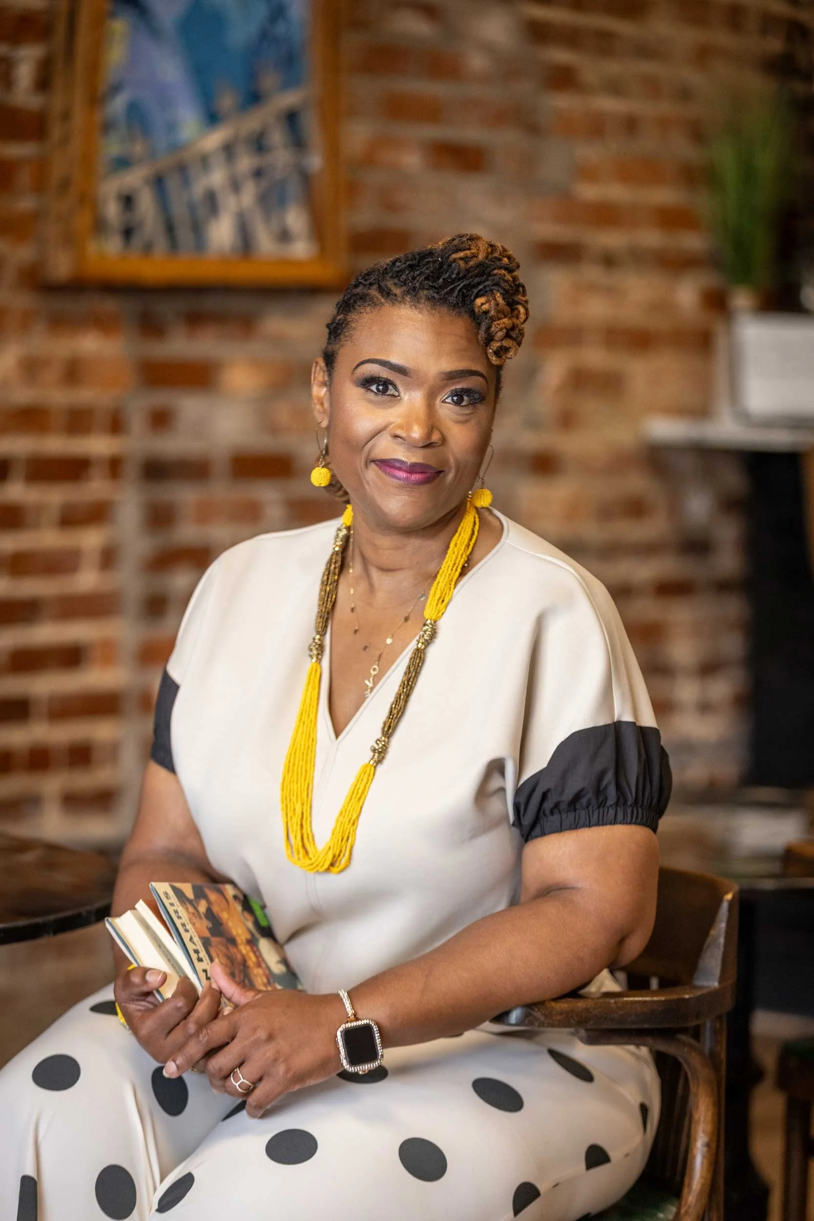 A woman with short, styled hair, wearing a cream-colored dress with black sleeves and black polka dots, yellow jewelry, and a smart watch, sitting in a space with a brick wall and artwork in the background, holding a small booklet.