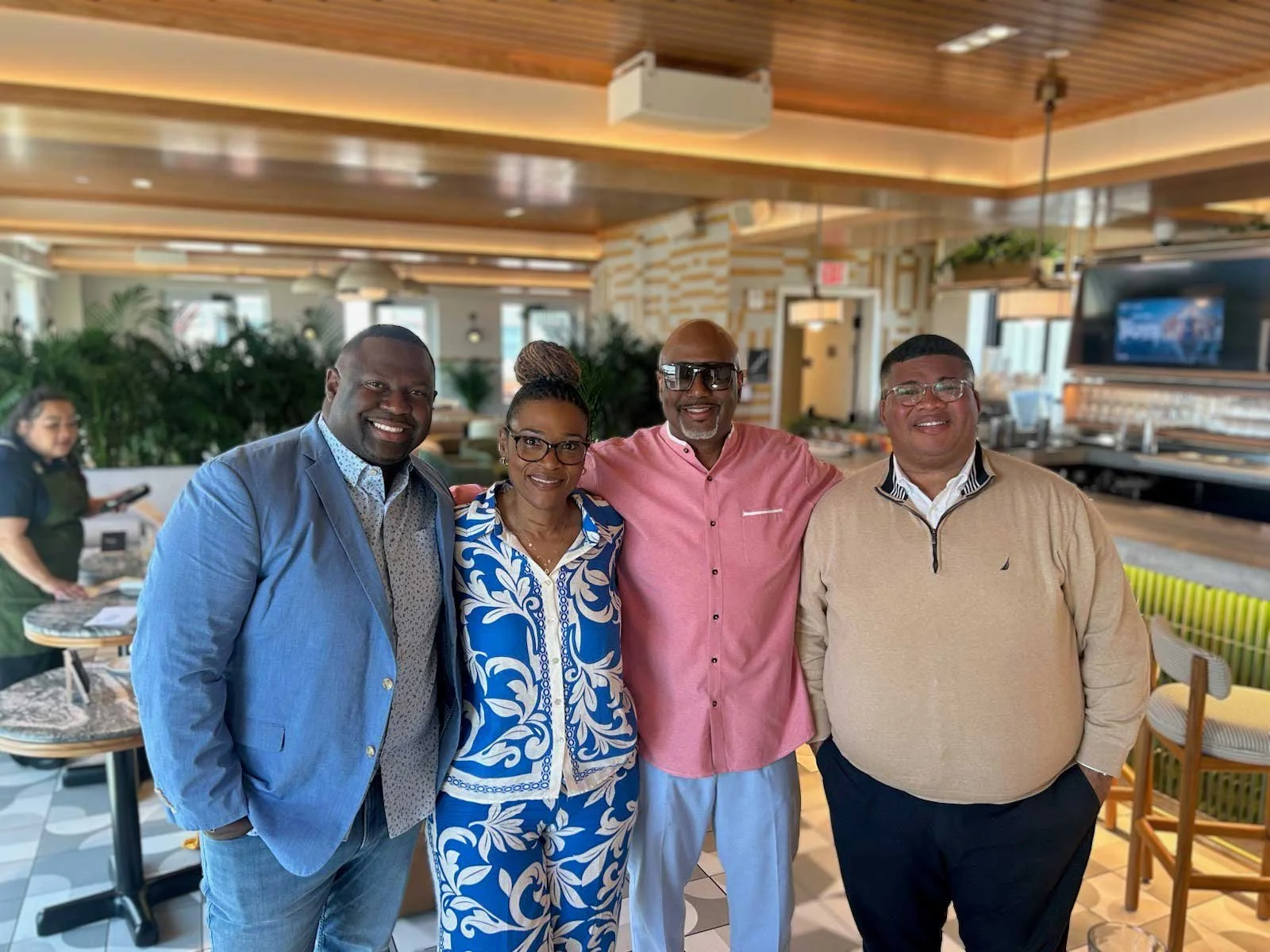 Four people standing closely together, smiling, inside a well-lit restaurant or cafe with wooden ceiling and greenery in the background.