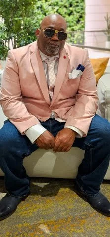 A man wearing a light pink blazer, sunglasses, and a white shirt sitting on a beige couch outdoors, with greenery and a pink building in the background.