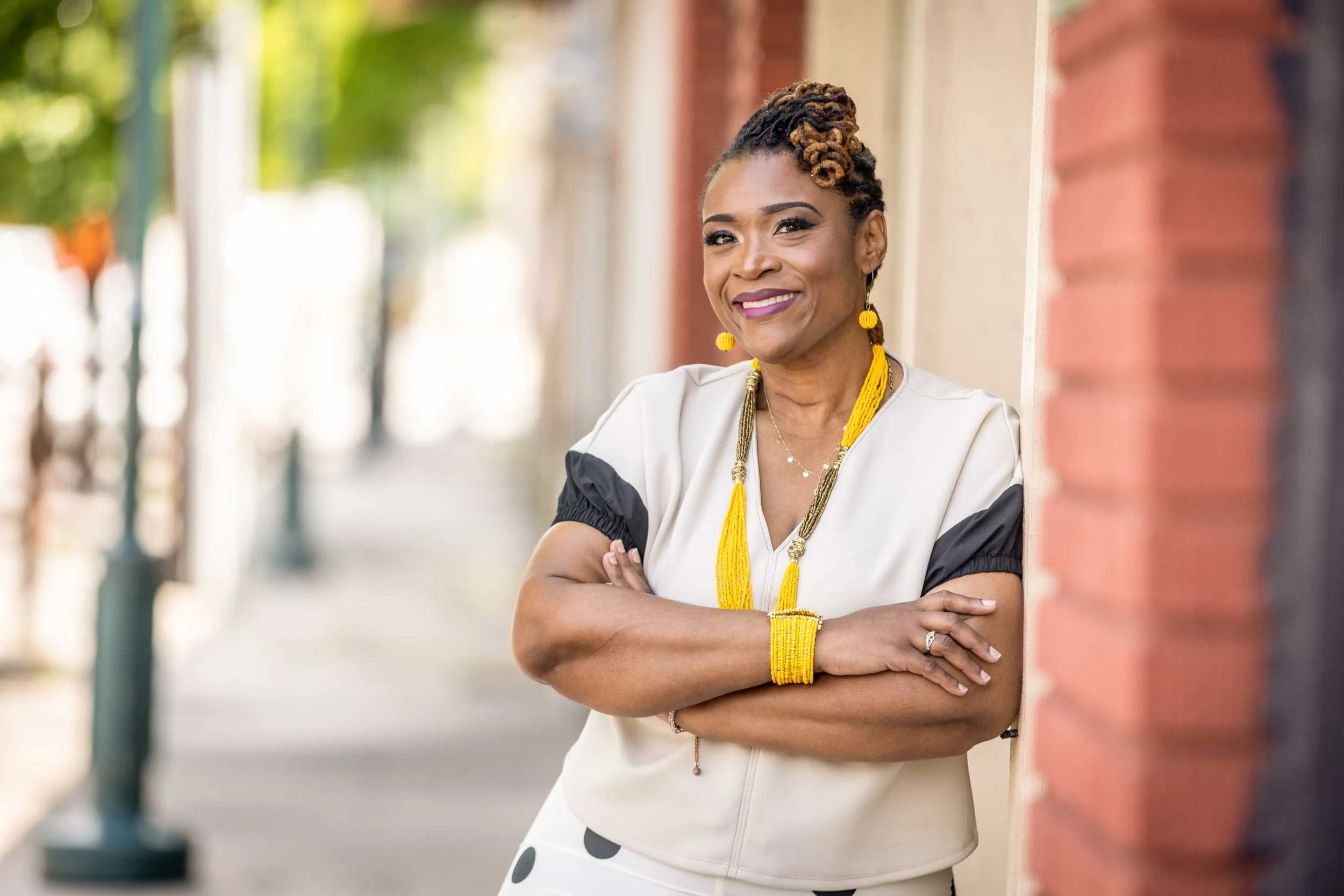 A woman with short dreadlocks styled in an updo, smiling with arms crossed, standing outside next to a brick wall with a blurred background of trees and a sidewalk.