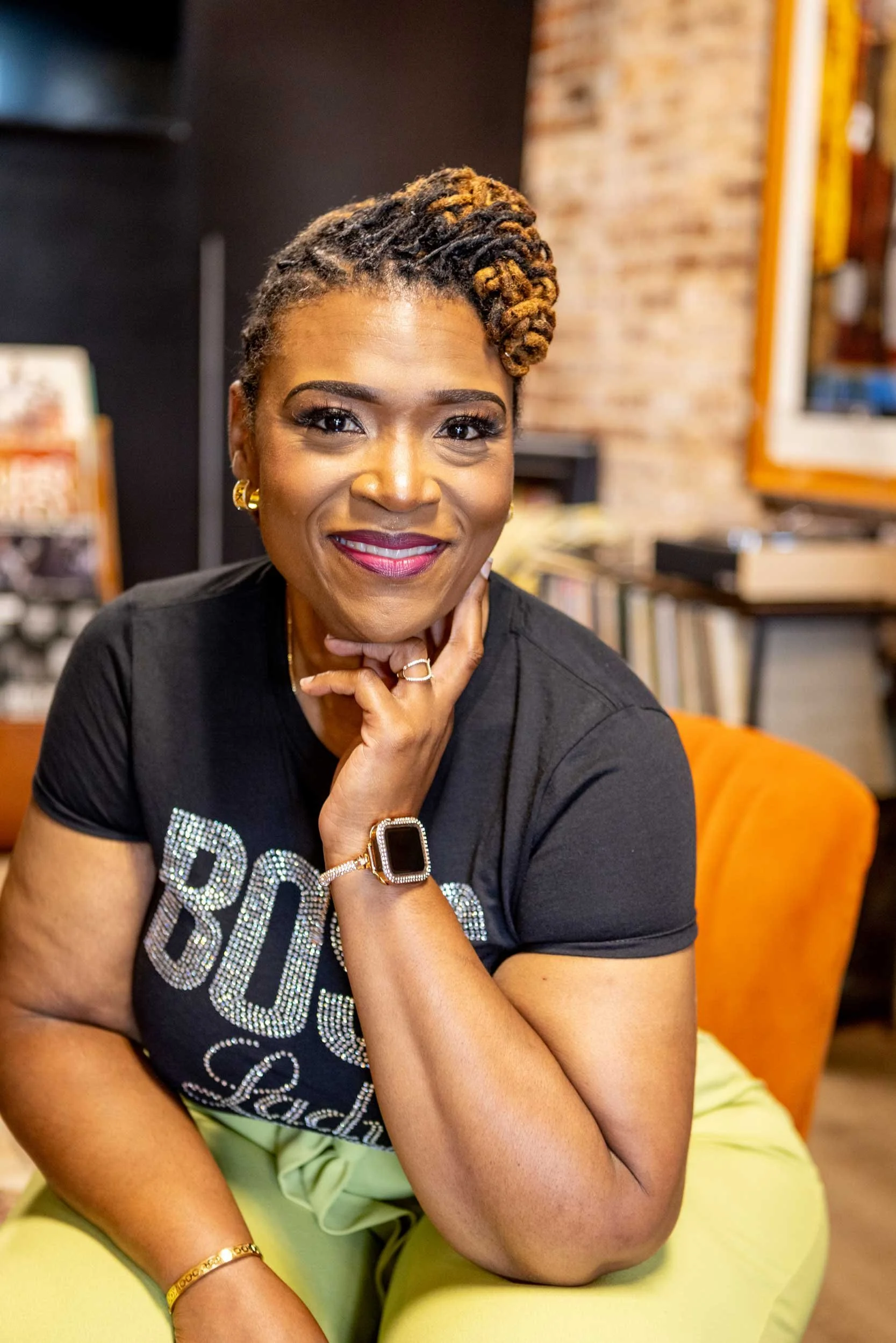 A smiling woman with short, twisted, highlighted hair, wearing a black T-shirt with rhinestone lettering, gold jewelry, and sitting on an orange couch in a room with a brick wall background.