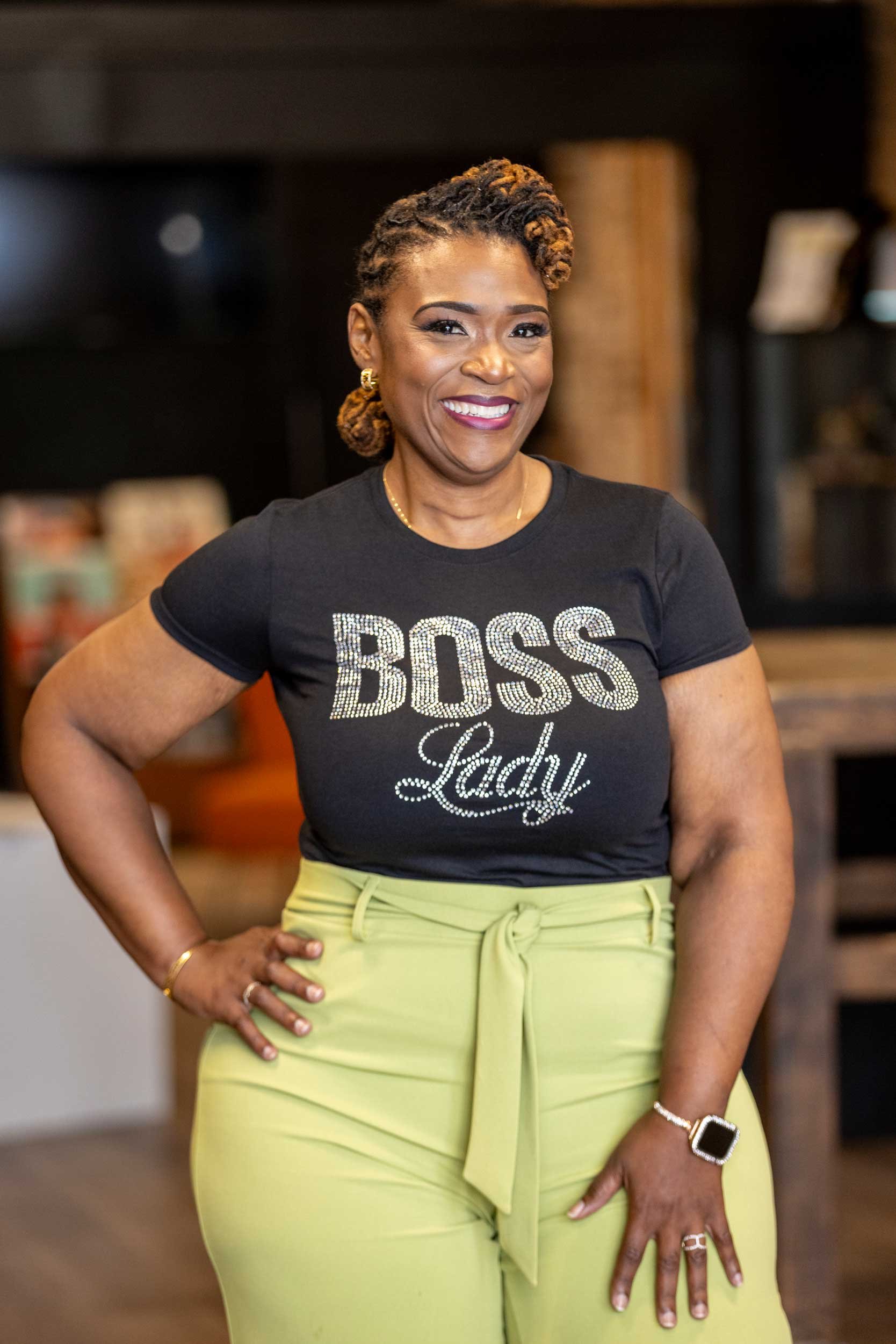 A woman smiling, wearing a black T-shirt with the words 'BOSS Lady' in sparkling letters, yellow high-waisted pants, and jewelry, standing indoors with a blurred background.