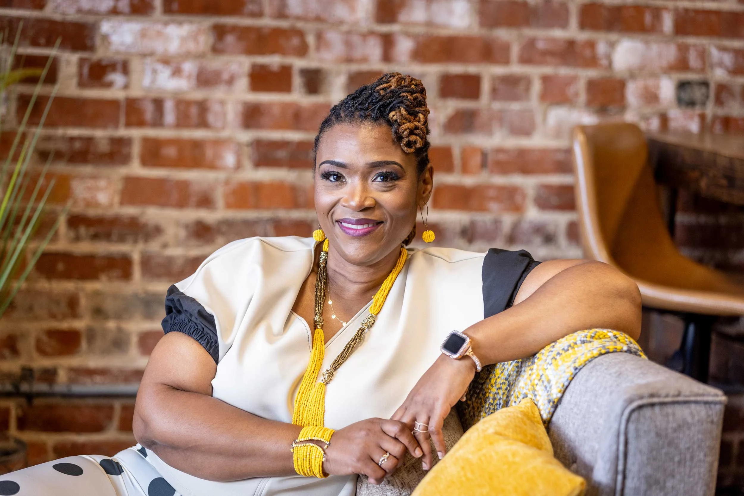 A woman smiling with dreadlocks styled up, wearing yellow jewelry and a beige top, sitting in front of a brick wall.