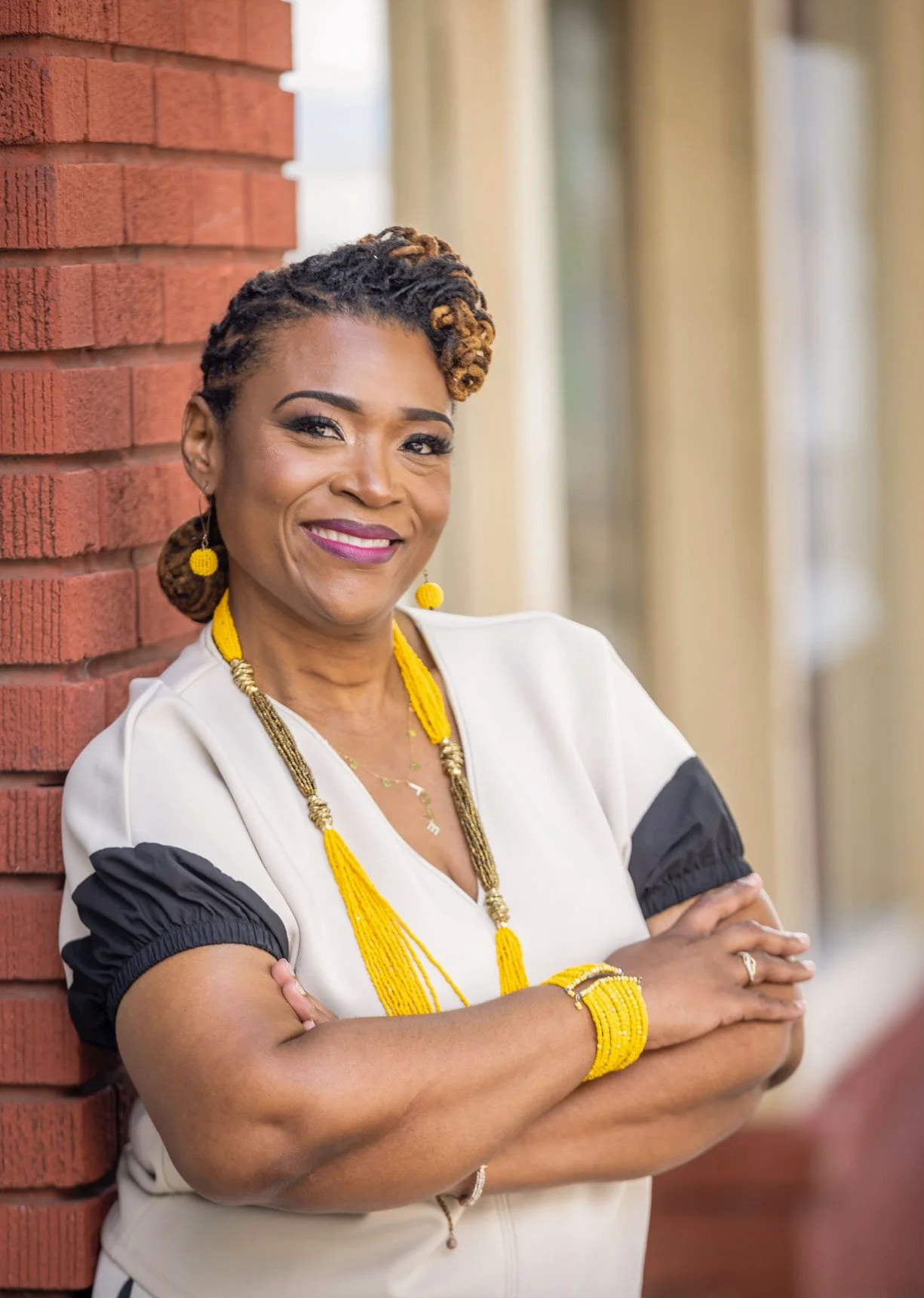 A confident woman with natural curly hair, wearing a white top with black accents, accessorized with yellow jewelry, standing with arms crossed beside a red brick wall.