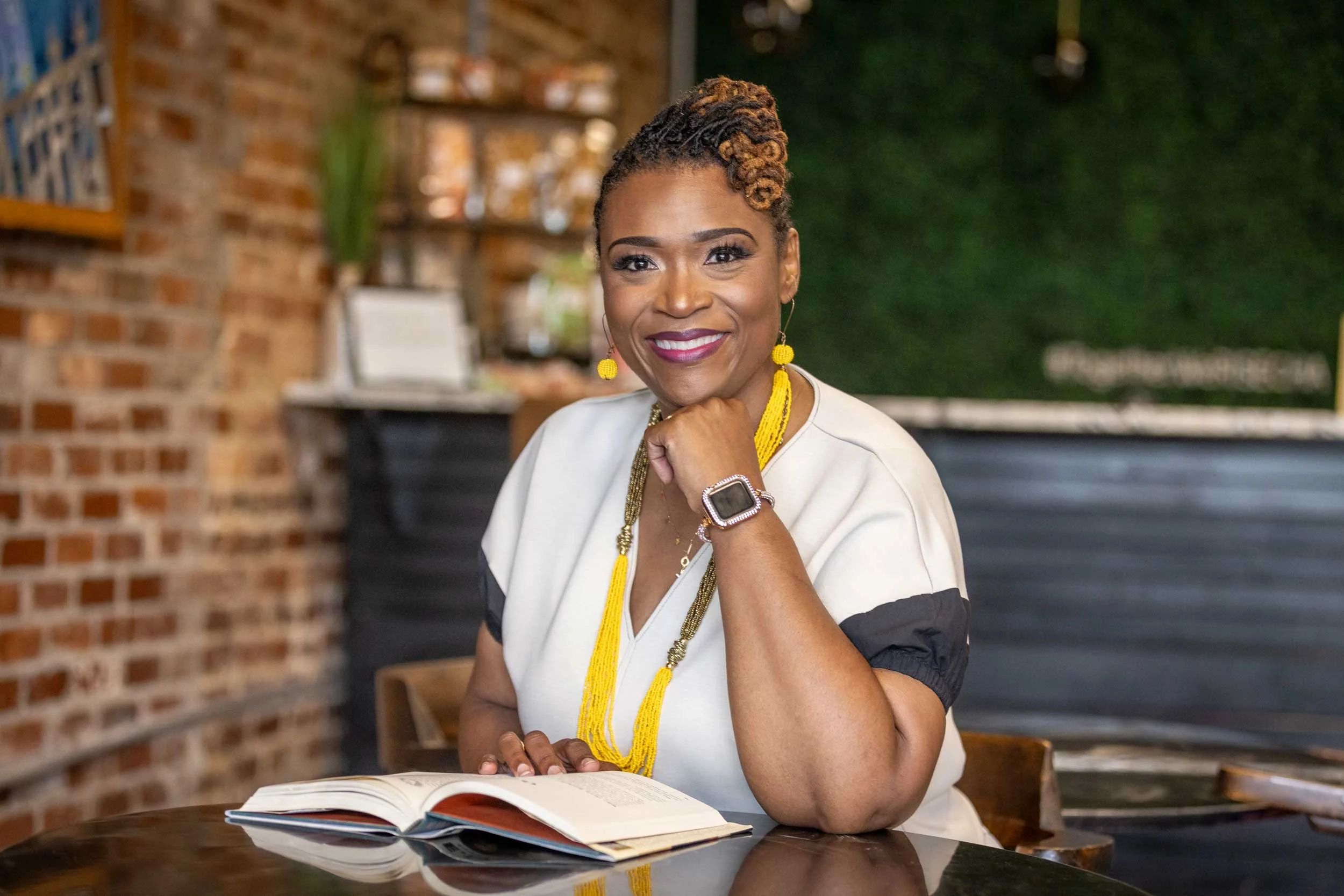 A woman with short curled hair, wearing yellow jewelry, sitting at a table with an open book, posing with her chin resting on her hand and smiling.