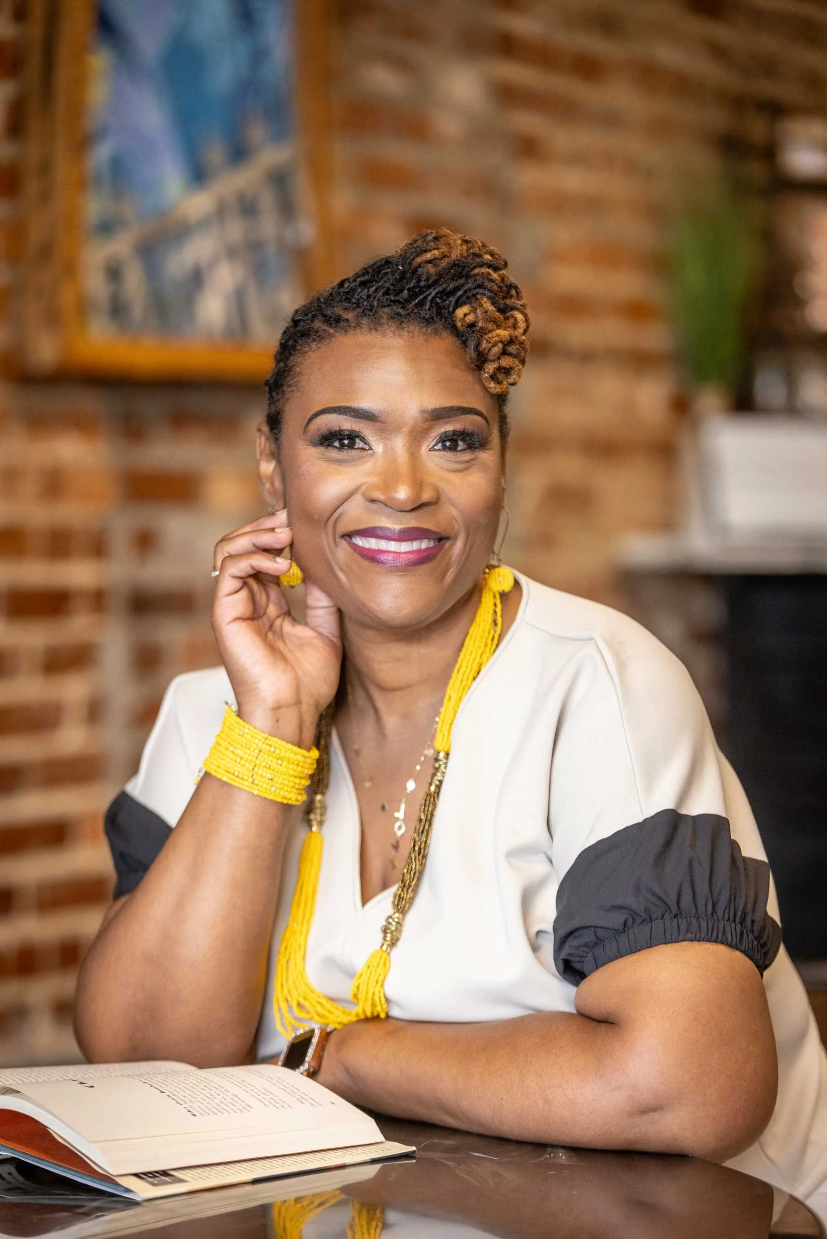 A smiling woman with short curled hair, wearing a cream-colored top with black sleeves, yellow jewelry, and a yellow necklace, sitting at a wooden table with an open book, in a setting with a brick wall background and artwork in the background.