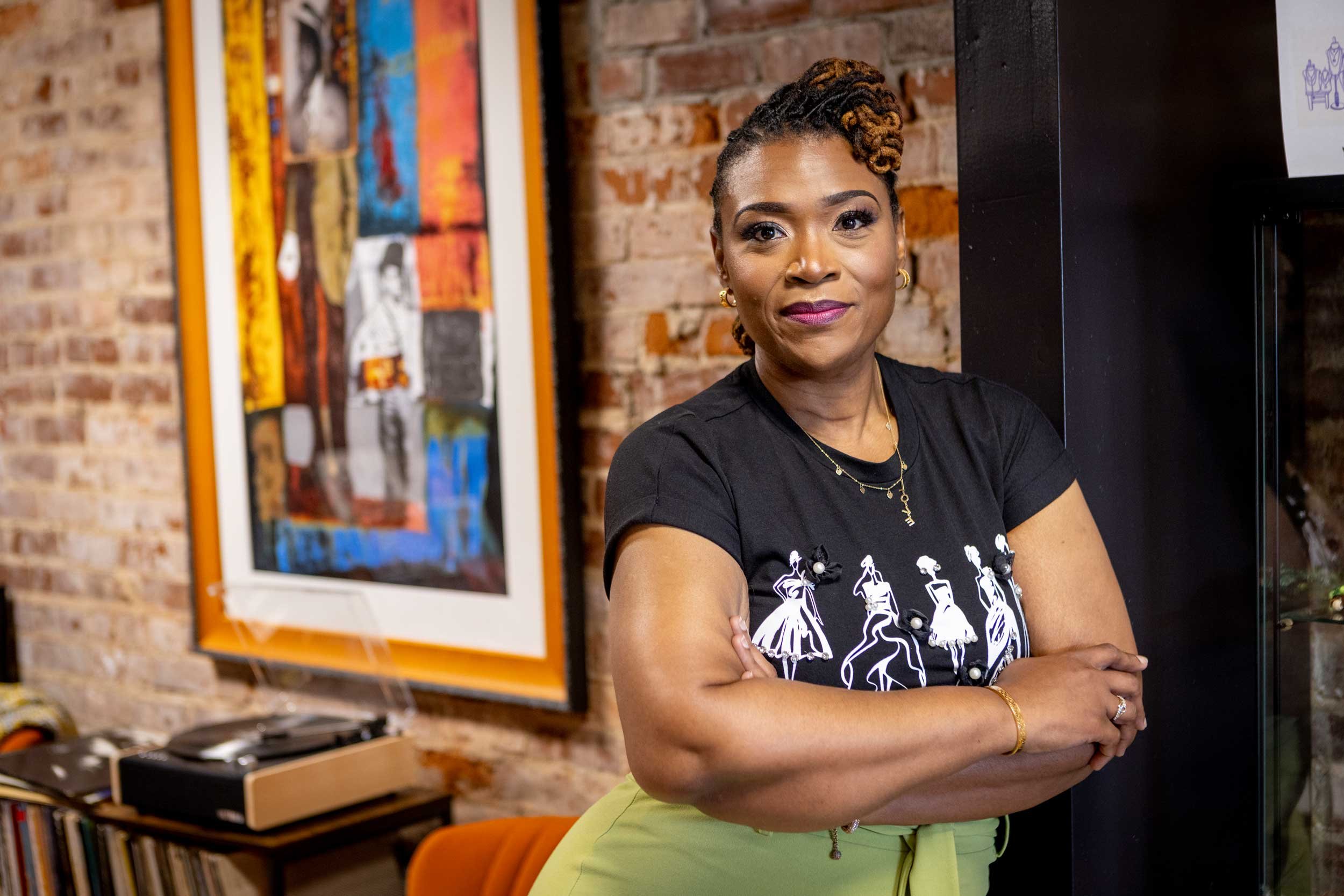 A woman with braided hair and earrings, wearing a black t-shirt and lime green skirt, standing with arms crossed in a room with a brick wall and colorful artwork.
