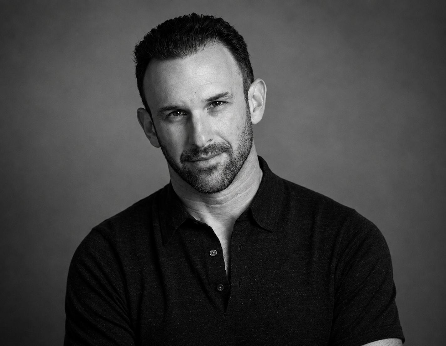 Black and white portrait of a man with short dark hair and a beard, looking at the camera with a slight smile, wearing a dark collared shirt against a plain background.