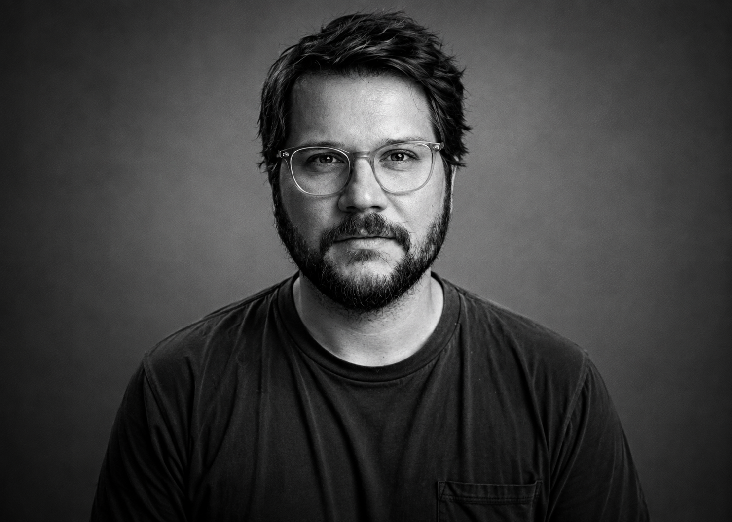 Black and white portrait of a man with glasses, a beard, and tousled hair, wearing a dark t-shirt, looking directly at the camera against a plain background.
