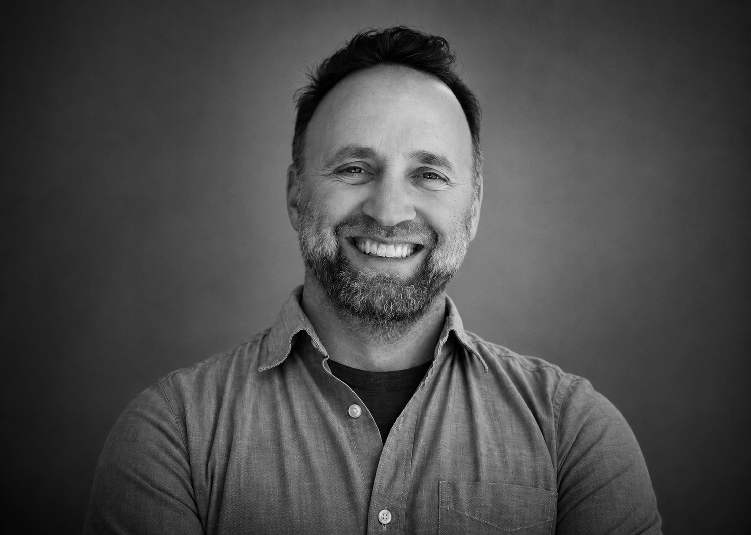 A black and white portrait of a smiling man with a beard and short hair, wearing a button-up shirt, in front of a plain gradient background.
