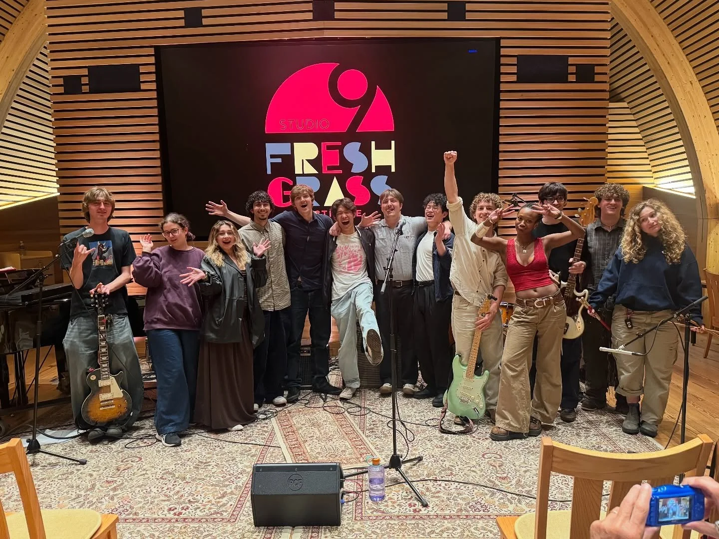 Group of people on stage in a music studio, holding instruments and celebrating, with a large sign reading 'Studio 9 Fresh Gas' in the background.
