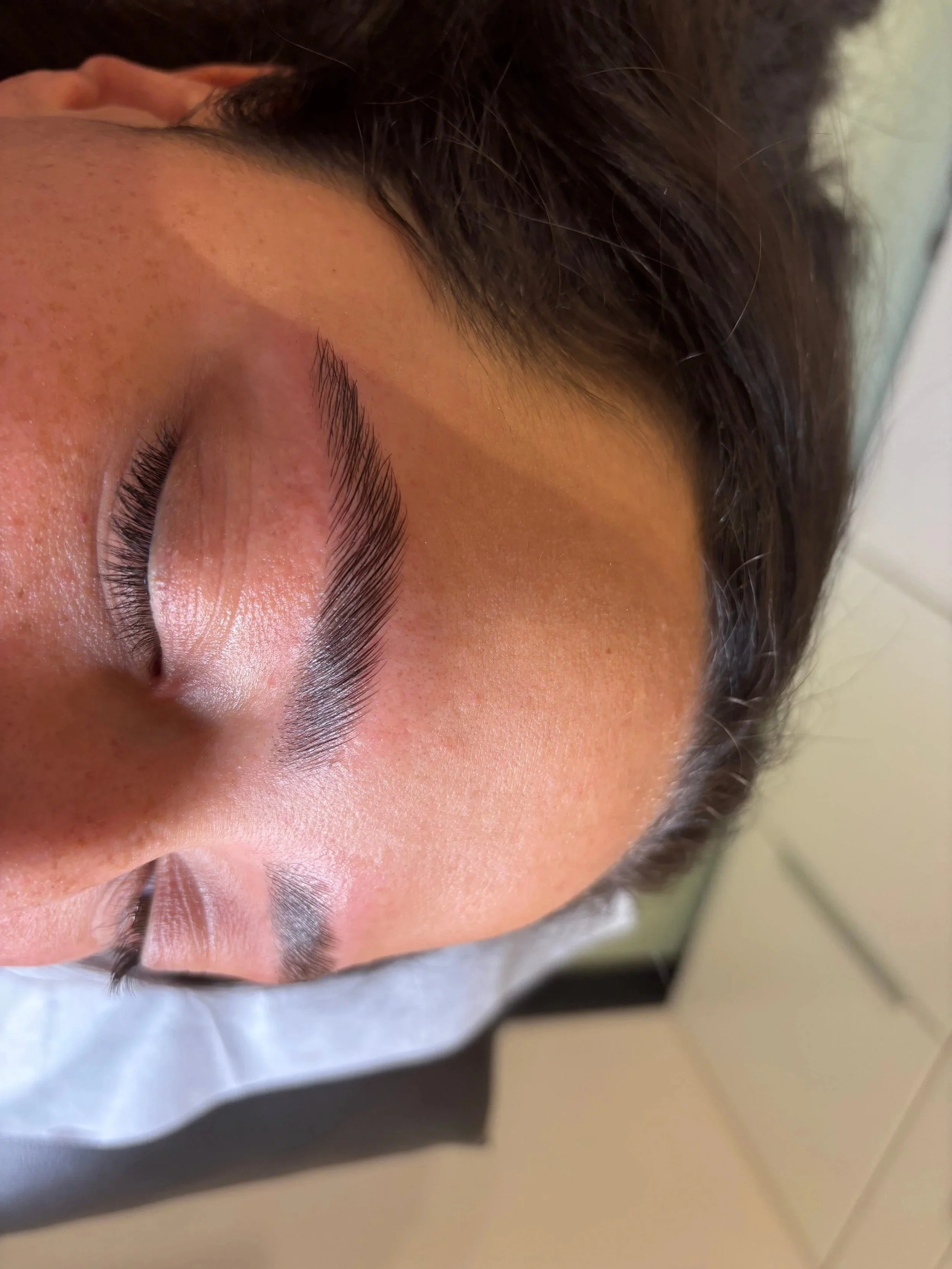 Close-up of a person's eye with long lashes, natural eyebrow, and skin texture, lying on a surface with part of the face covered by a dark cloth.