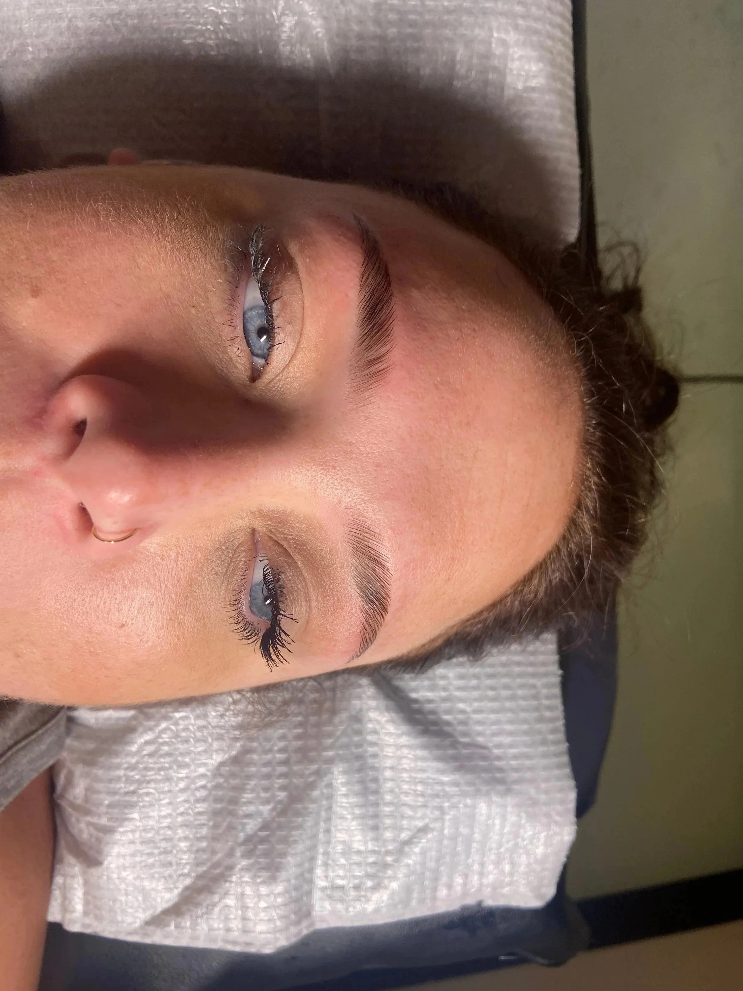 A person lying down with their eyes open, showing their face, eyebrows, and eyelashes. They have light skin, blonde eyebrows, and blue eyes. Their hair is curly and dark brown. They are wearing a gray shirt and a small nose ring.