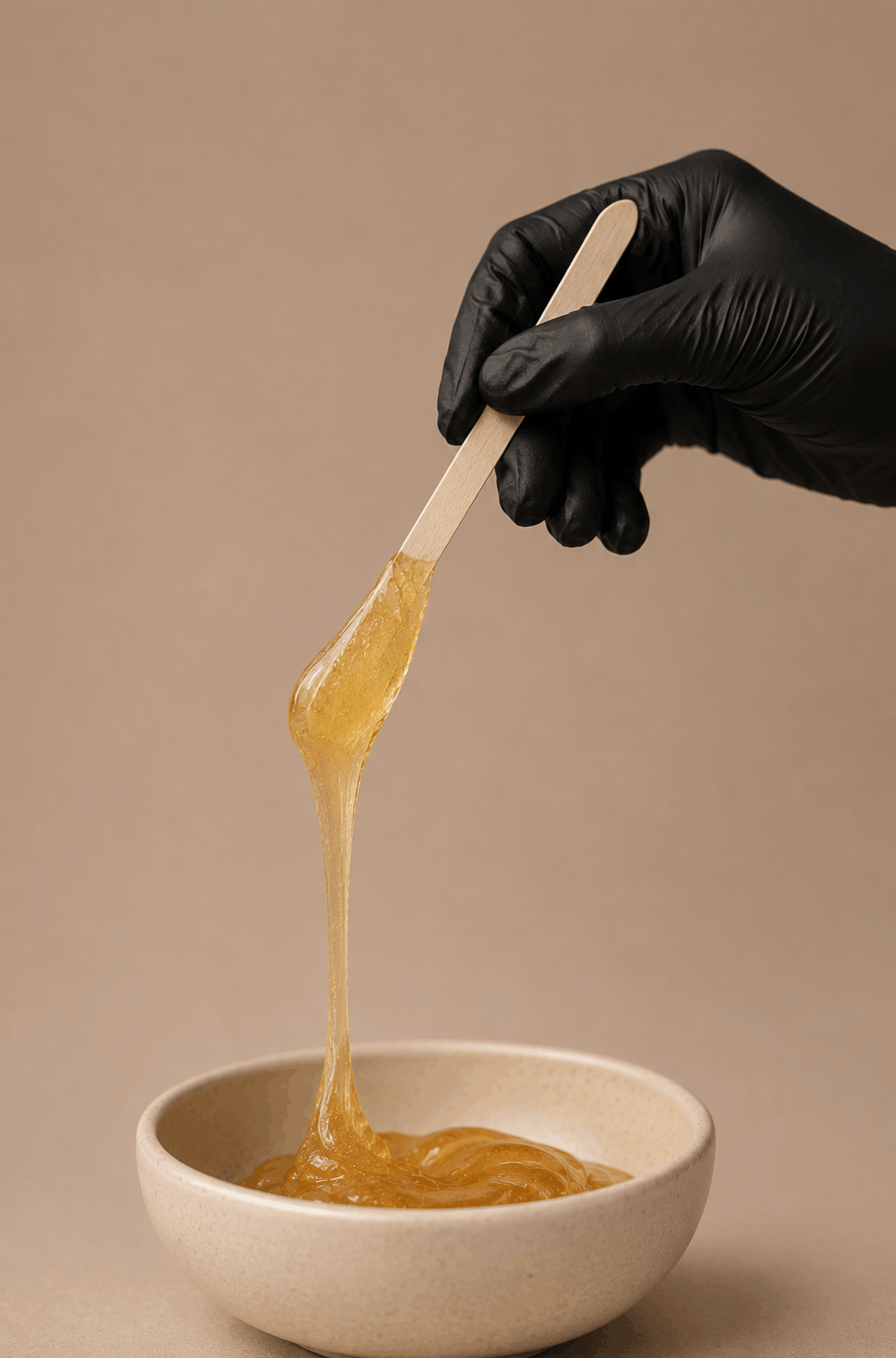 A person wearing a black glove holding a wooden spatula with sticky, golden substance over a beige bowl.