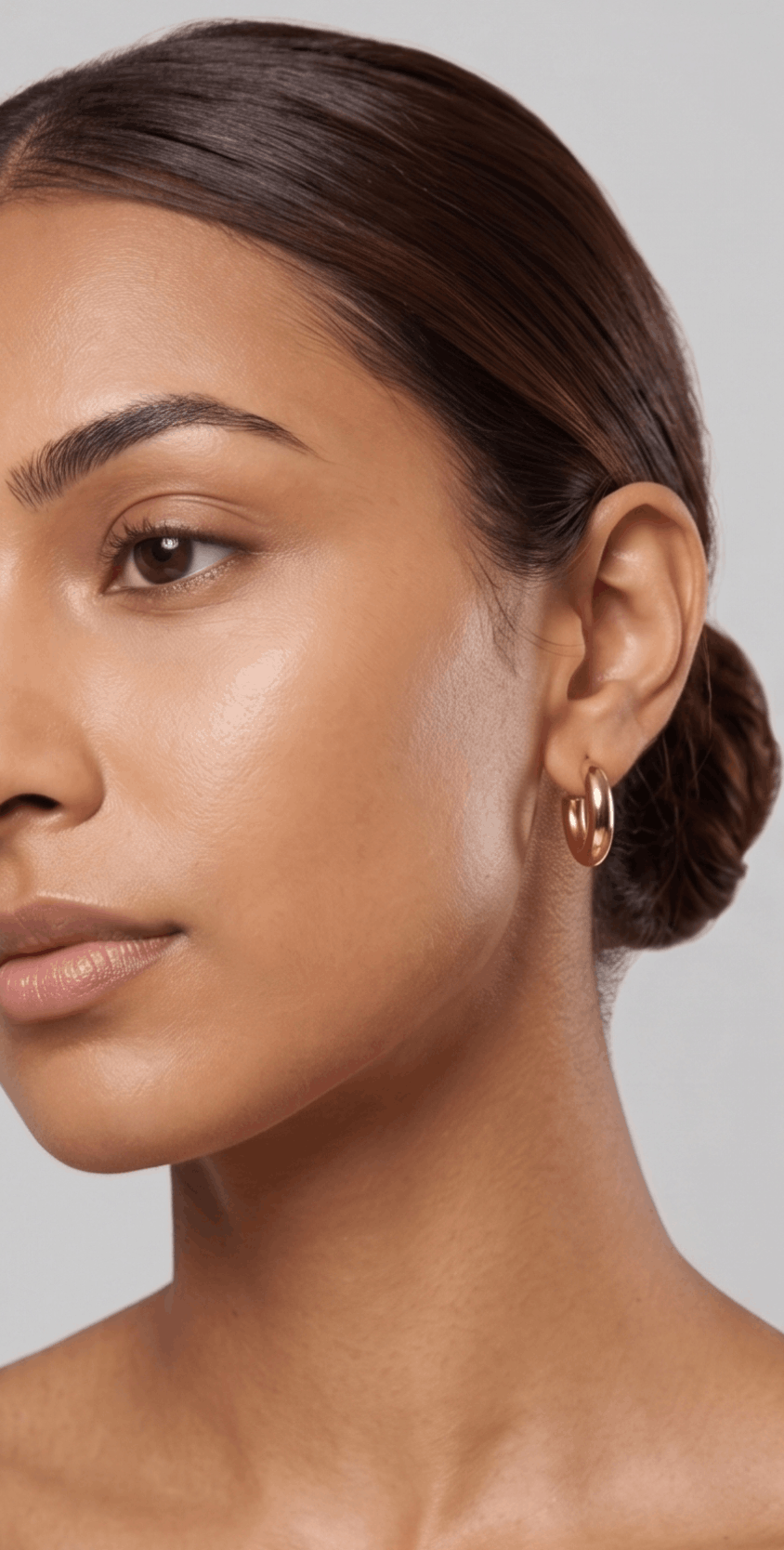 Close-up of a woman with smooth, glowing skin, wearing gold hoops earrings and a sleek, neatly styled bun, against a neutral background.