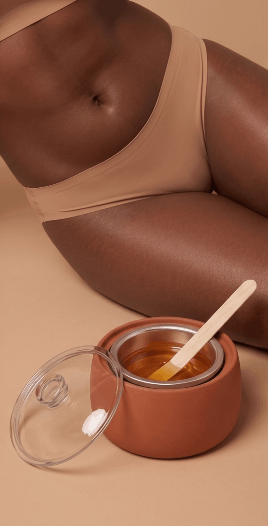 A person with dark skin wearing a beige tank top, sitting next to a pink ceramic wax warmer filled with honey wax, with a wooden spatula inside. The person’s torso and thighs are visible.