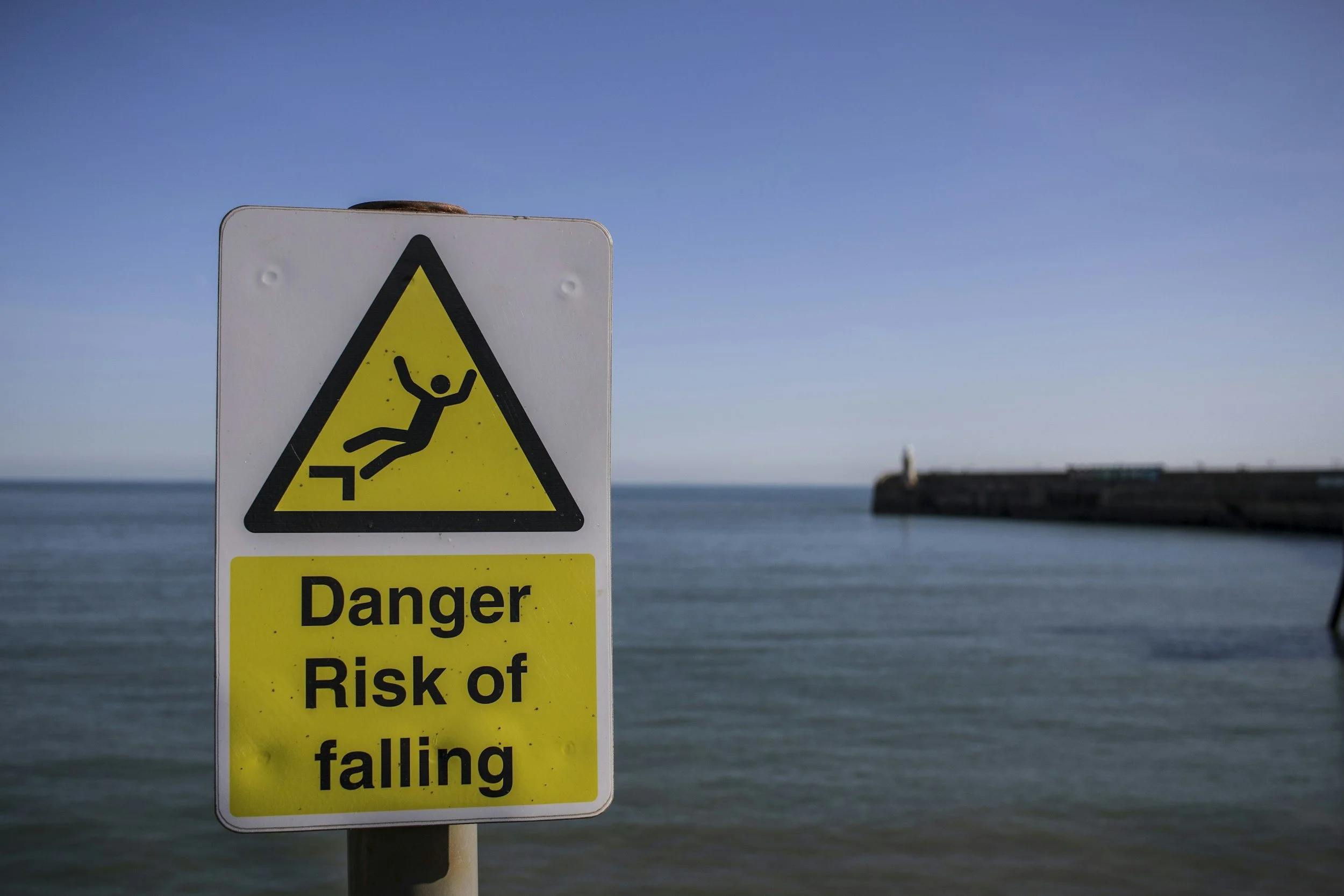 Warning sign with a yellow triangle and a stick figure falling, with a background of water and a distant pier