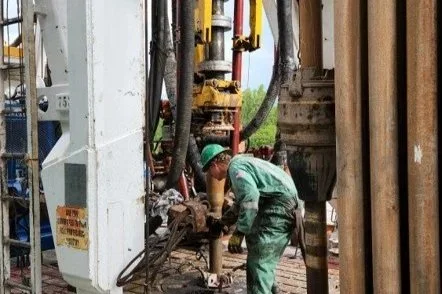 A worker inspecting or repairing equipment on a geothermal drilling rig, wearing a green uniform and helmet.