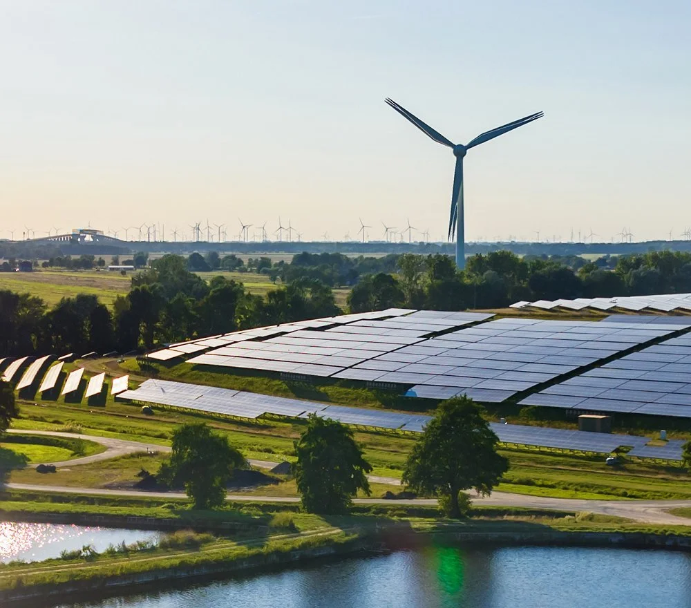 A landscape featuring a large wind turbine and solar panels, with trees, a small body of water, and a distant horizon with more wind turbines.