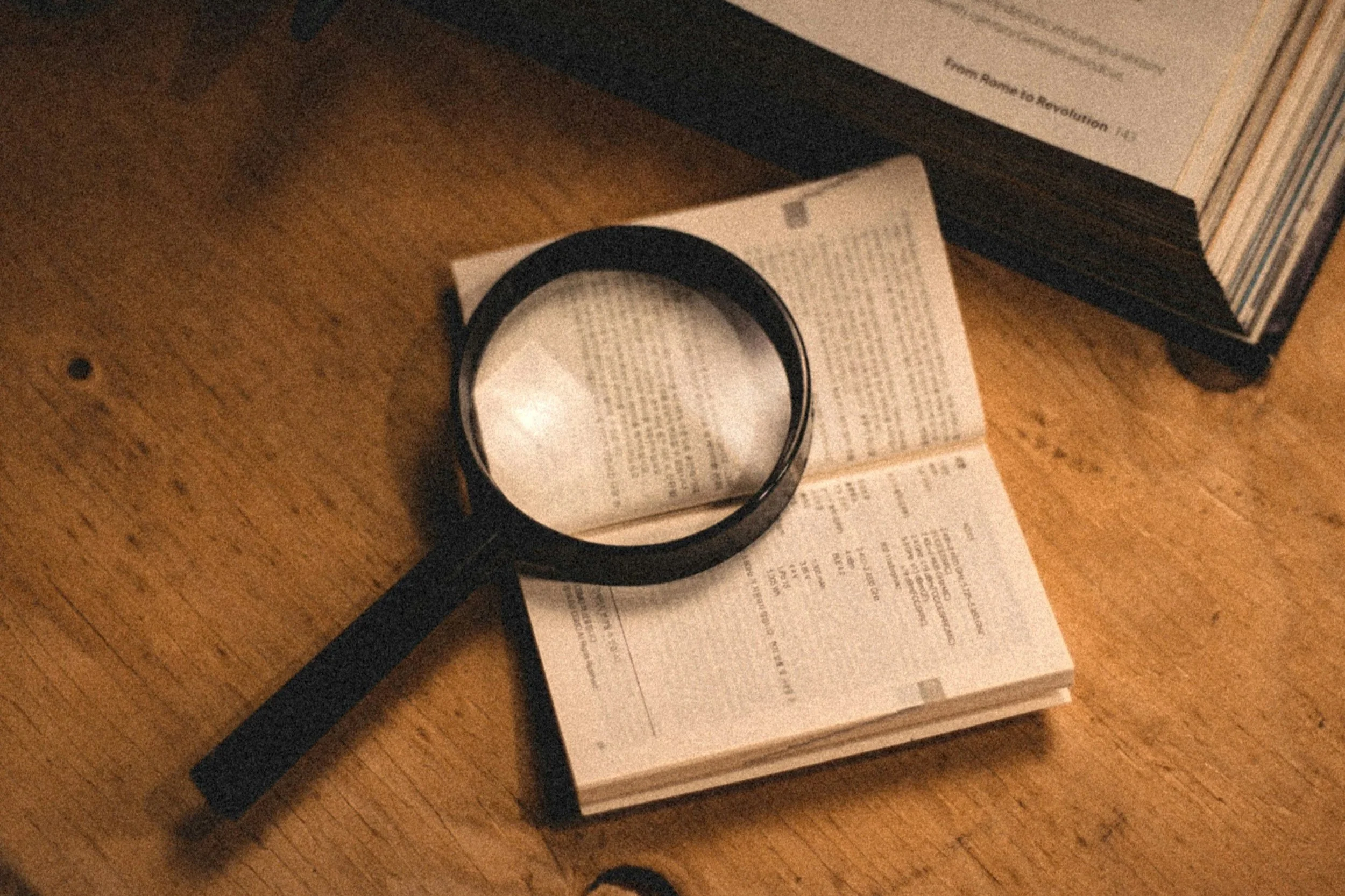Desk with a stack of books, a printed document, a black wristwatch or bracelet, and an open book on a wooden surface.