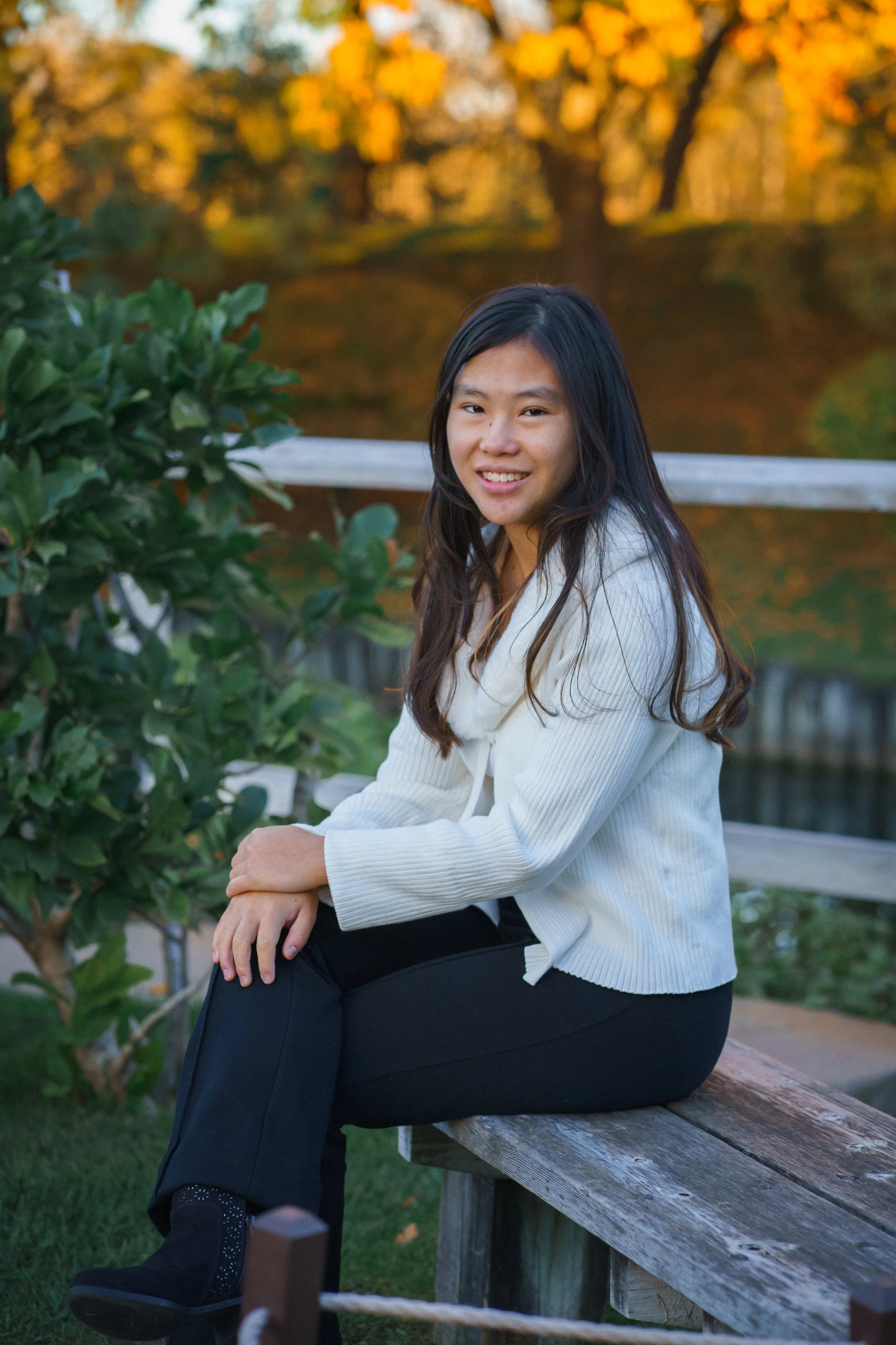 A young woman with long dark hair, wearing a white sweater and black pants, sitting on a wooden bench outdoors during fall. She is smiling at the camera with trees showing autumn foliage in the background.